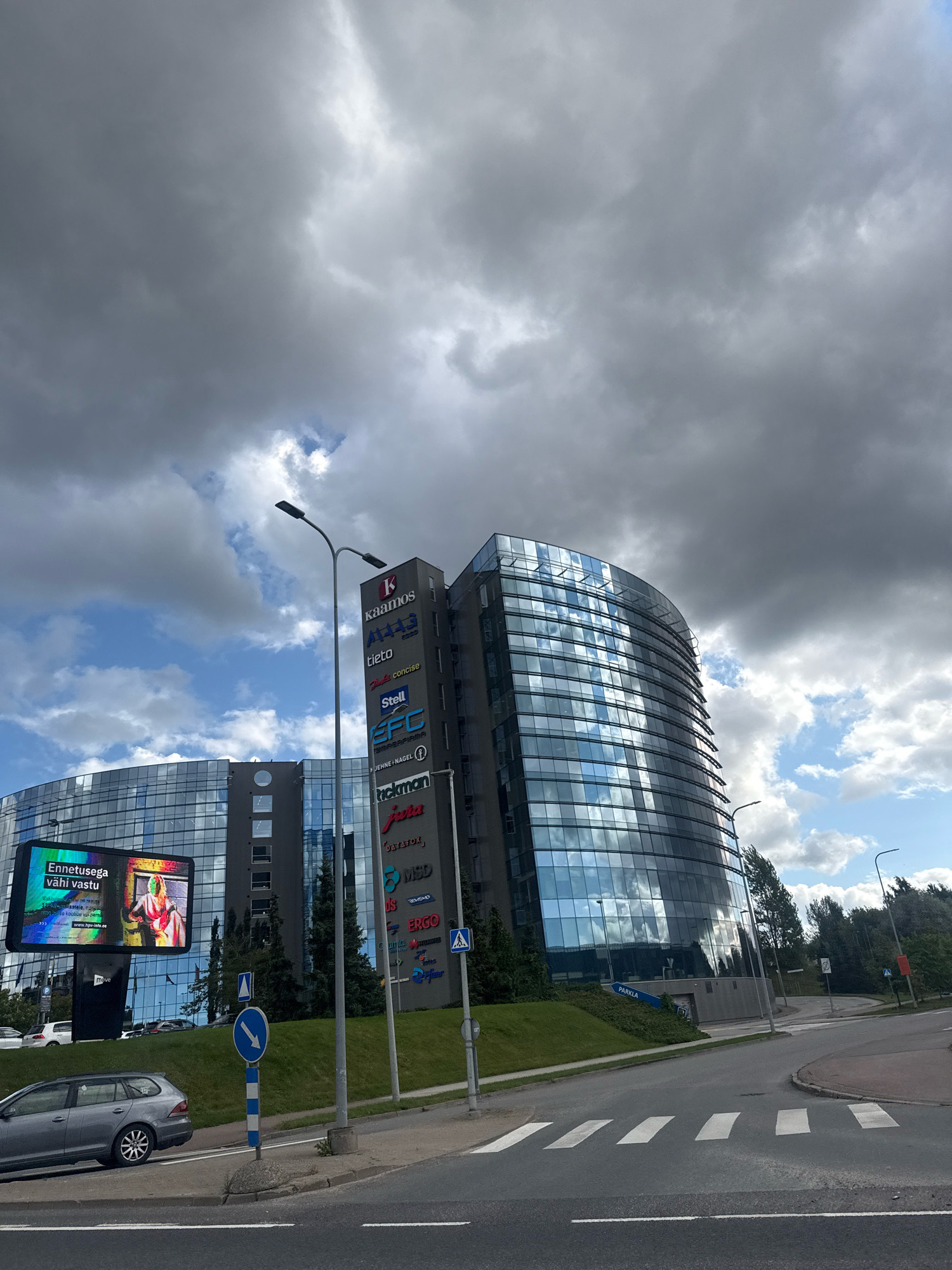 The image depicts a view of the modern office buildings of the Technopolis Ülemiste campus in Tallinn, Estonia. The buildings feature extensive glass facades that reflect the cloudy sky. A tall, slender tower with several company logos, including Kaamos, Ajakiri, Tieto, and Efcon, stands prominently. Adjacent to it, a curved building displays a large digital billboard with colorful graphics and text in Estonian that appears to be an advertisement. In the foreground, a grey station wagon is parked on the side of the road, and a pedestrian crossing is visible on the asphalt. The scene is set on a cloudy day, with dramatic clouds filling the upper portion of the frame. Trees and greenery surround the buildings. The overall impression is of a contemporary business and technology hub.