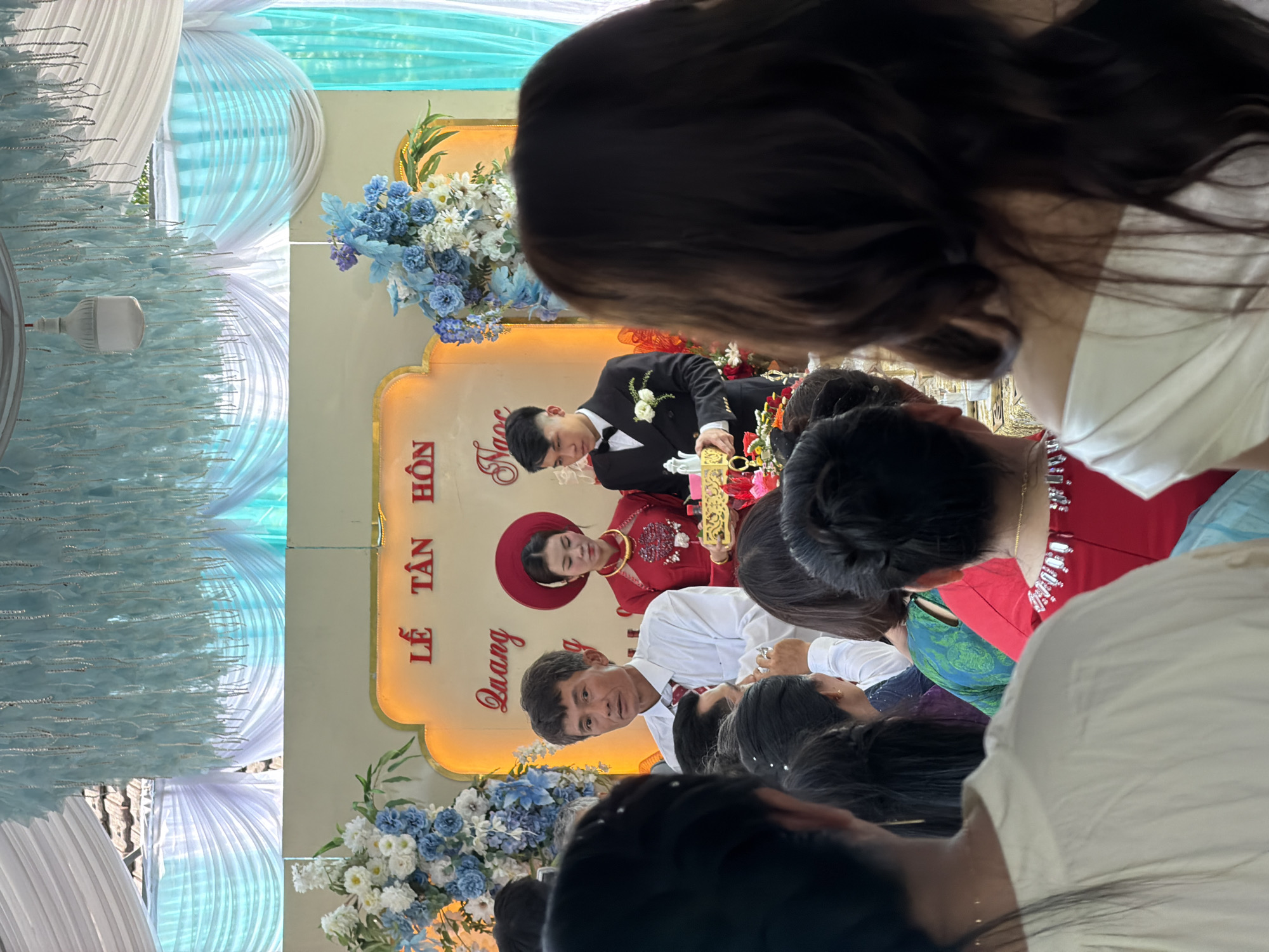 The image depicts an indoor wedding or nuptial ceremony ("Lễ Tân Hôn") taking place in Bình An, Vietnam. The scene is festively decorated with white and light blue drapes creating a soft background, interspersed with a stringy, feathery light blue decoration on the left. A white LED light bulb is visible hanging on the left side amidst these decorations.
In the center, against a cream-colored, ornate banner with gold trim, are the bride and groom. The bride is on the right, wearing a traditional red Vietnamese áo dài and a matching red non quai thao (conical hat). The groom is on the left, dressed in a black suit with a white shirt and bowtie. They are both holding a decorative golden, ornate box or item.
Floral arrangements featuring white, light blue, and purple flowers (likely hydrangeas and daisies) adorn the banner on both sides of the couple.
In the foreground and midground, numerous guests are present, mostly visible from behind, watching the ceremony. A man in a white shirt and dark pants is clearly visible in the midground, looking towards the couple. A woman in a green and blue patterned top is also partially visible from behind. The general atmosphere appears celebratory and focused on the couple.
Visible text on the central banner, written vertically in red, includes: "LỄ TÂN HÔN" (Nuptial Ceremony or Wedding Ceremony). Below this, also vertically, are two names: "Quang" and "Nước", likely representing the names of the groom and bride. The lighting is bright, suggesting either daytime or a well-lit indoor evening event.