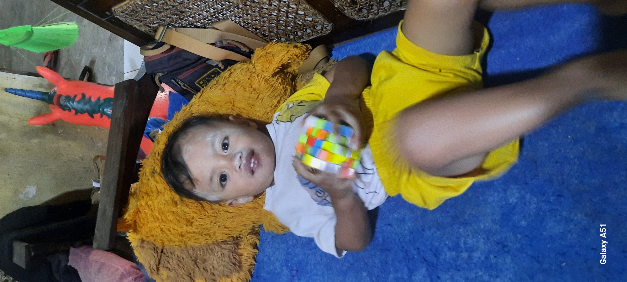 In a room in Ploso, Indonesia, a young child lies on a blue carpet, resting against a large, fluffy yellow stuffed animal. The child, with white powder on their forehead and cheeks, wears a white t-shirt and yellow shorts. They are holding a colorful Rubik's Cube in their hands. The background shows a wall, a wooden plank, and a woven chair. A red and blue inflatable toy with a horn is visible behind the child.