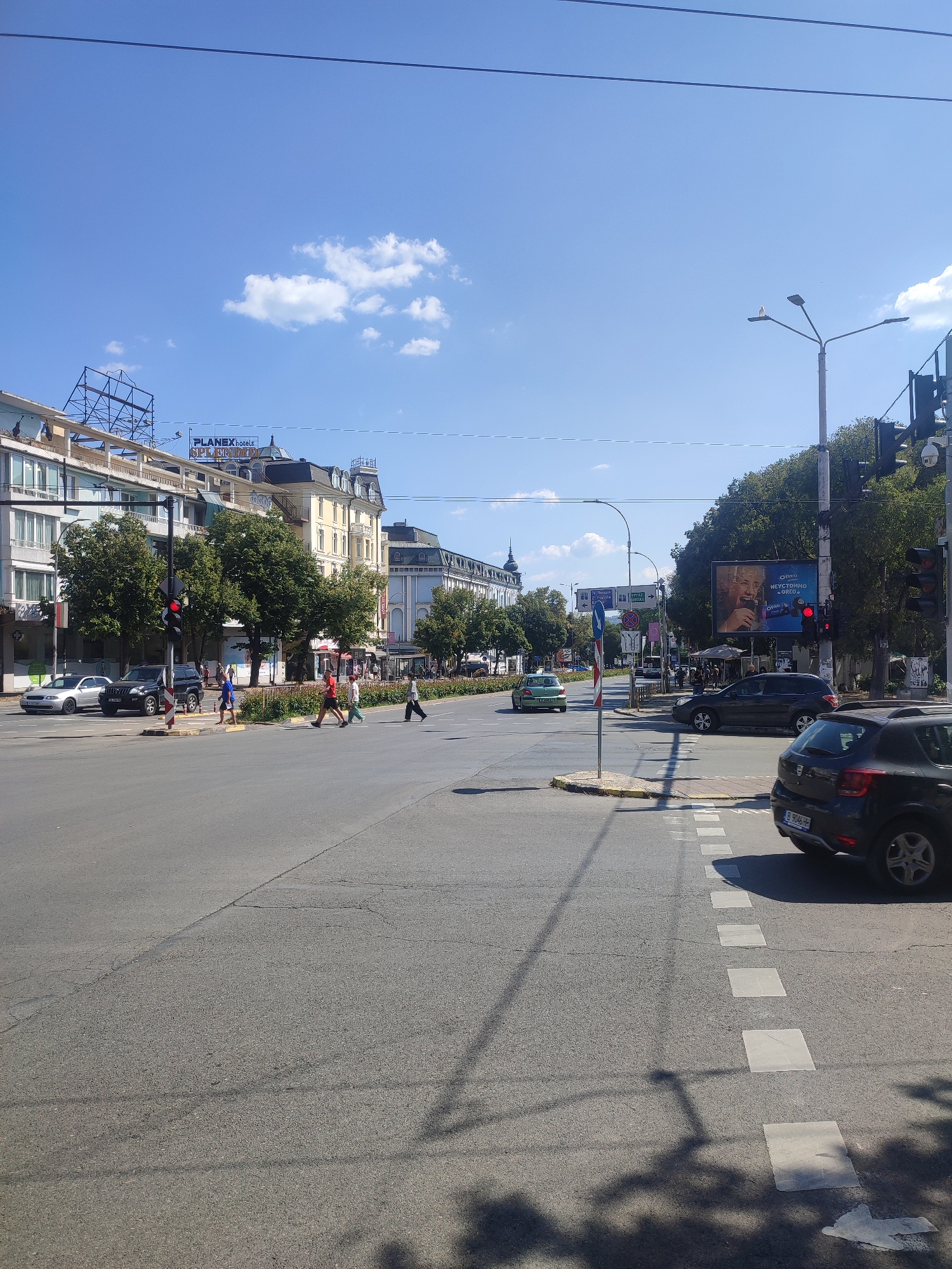 This is a daytime, eye-level, medium-shot photograph of a city intersection in Varna, Bulgaria. The sky is clear blue with a few scattered white clouds. The foreground features an asphalt road with white dashed lines and square pedestrian crossing markings. Several cars are visible, including a dark-colored SUV and a silver sedan in the left lane. On the right side of the road, a dark gray hatchback is turning.
In the middle ground, a traffic light shows red. Pedestrians are crossing the street at the marked intersection. To the left of the intersection, there are several multi-story buildings with signs for "PLANEX hotels" and "SPLENDID". Trees with green foliage line the sidewalk in front of these buildings.
In the background, more buildings are visible, including some with ornate architecture and a building with a domed roof. A large billboard advertising Oreo is also visible on the right side of the intersection. Streetlights and traffic signals are present throughout the scene. The overall environment suggests a busy urban setting on a sunny day.