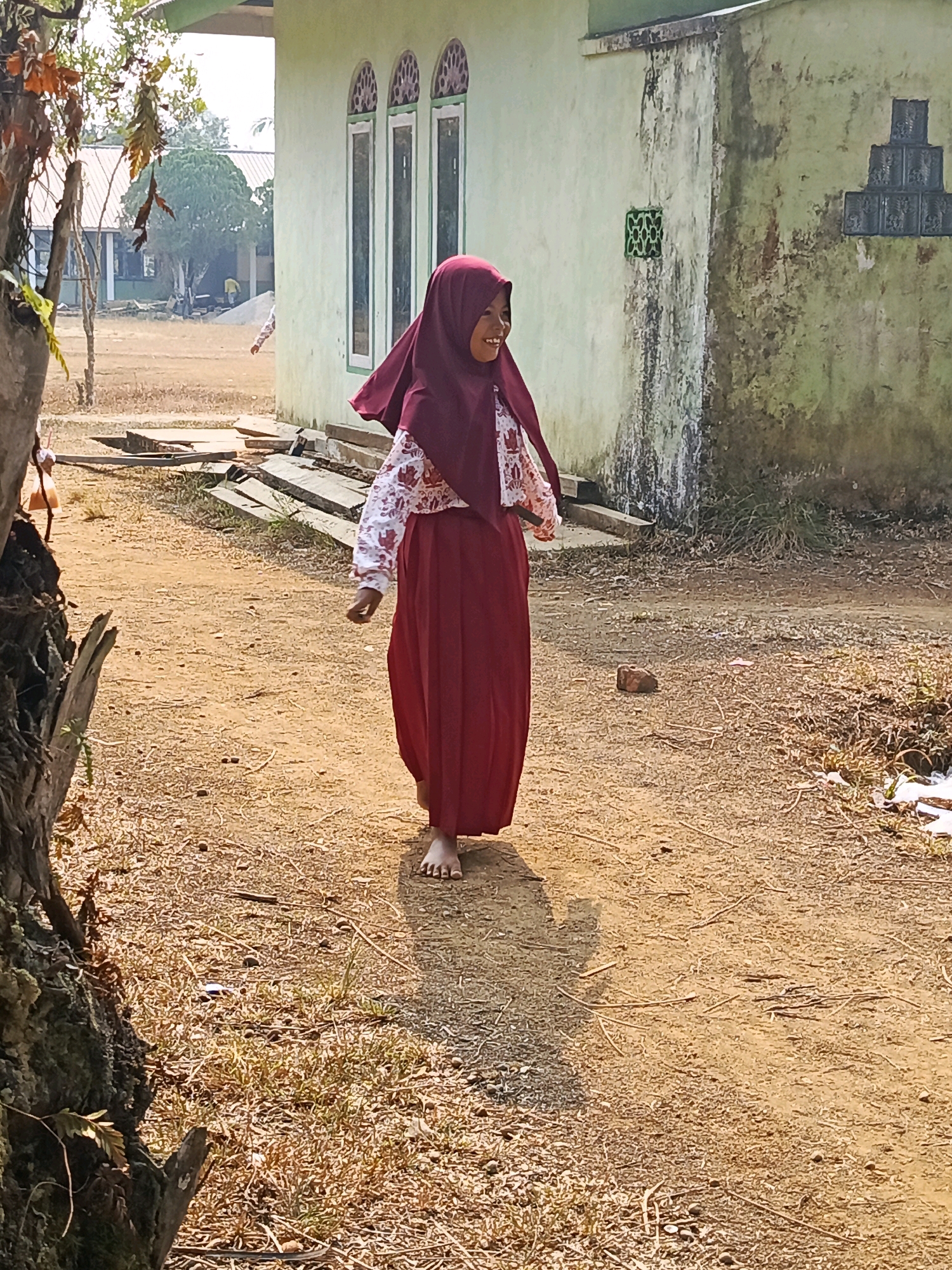 A young girl wearing a maroon hijab and a red skirt walks barefoot on a dirt path in front of a light green building. She has a white shirt with a floral pattern underneath her hijab and over her skirt. The building behind her has three arched windows with decorative patterns and a section of textured wall with a geometric pattern above it. The ground is dry and dusty, with scattered dry grass and twigs. In the background, there are trees and other structures, including a building with a white roof. The lighting suggests it is daytime, with shadows cast by the girl and objects. The overall scene appears to be in a rural or semi-rural area.