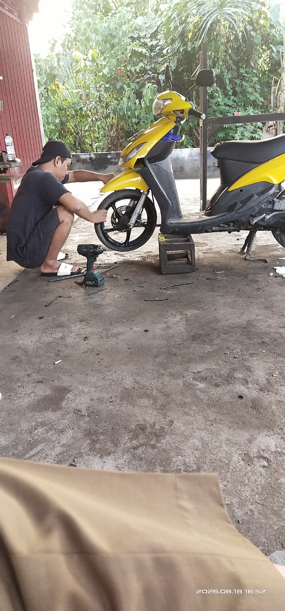 In Matabu, Indonesia, a man wearing a dark t-shirt and shorts, along with a black baseball cap, is crouched on the ground, working on the front wheel of a yellow and black scooter. The scooter is elevated by wooden blocks. A green electric impact wrench and various tools are scattered on the concrete floor around him. The scene appears to be an outdoor workshop or a repair area, with lush green foliage in the background and a red corrugated metal wall to the left. The ground is dusty concrete, showing some tire marks and debris. A bottle of water and a rag are visible on a shelf in the background. A tan fabric occupies the bottom foreground of the image. The timestamp "2026.08.18 16:52" is visible in the bottom right corner.