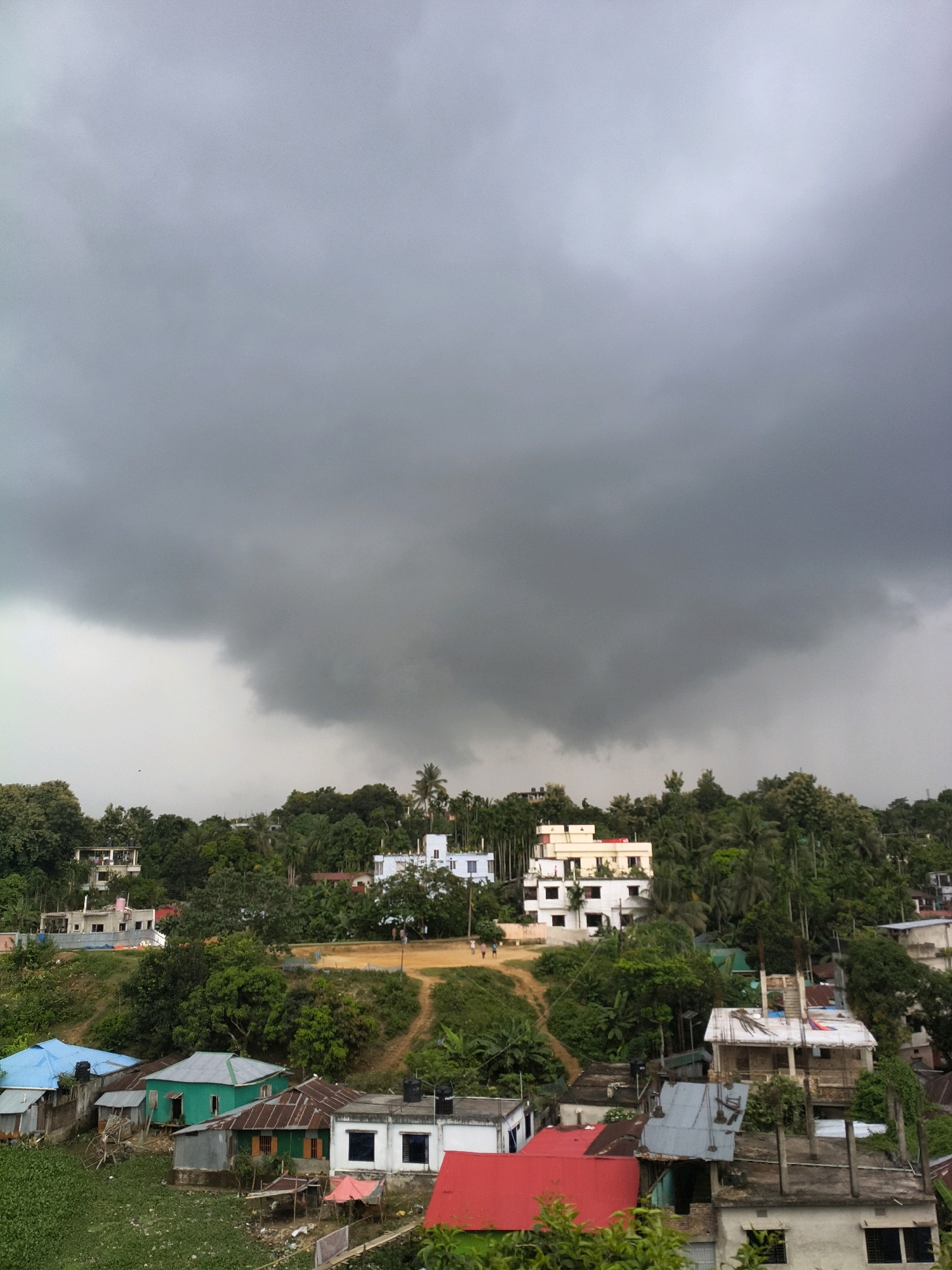An elevated, wide shot captures a residential area in Rangamati, Bangladesh, under a heavily overcast sky that portends rain. A large, dark grey cloud dominates the upper half of the frame, obscuring the sky. Below this imposing cloud, a verdant hillside is densely populated with trees and a mix of modest houses and larger, multi-story buildings.
In the foreground, several houses with corrugated metal roofs are visible. One prominent house has a bright green wall and a weathered metal roof. Adjacent to it is a white house with blue-tinted windows. Further to the left, a house is covered with a blue tarp roof. Scattered debris and some vegetation can be seen around these homes.
The middle ground features more homes, including a striking white building with multiple floors and balconies, and a cream-colored building with a red-tiled roof. A dirt path winds up the hillside between the houses, with a few figures walking along it.
The background is filled with lush green trees and foliage, creating a layered landscape. In the distance, more houses are interspersed among the trees, suggesting the dense nature of the settlement. The overall impression is one of a community nestled within a natural, hilly environment, awaiting a storm.