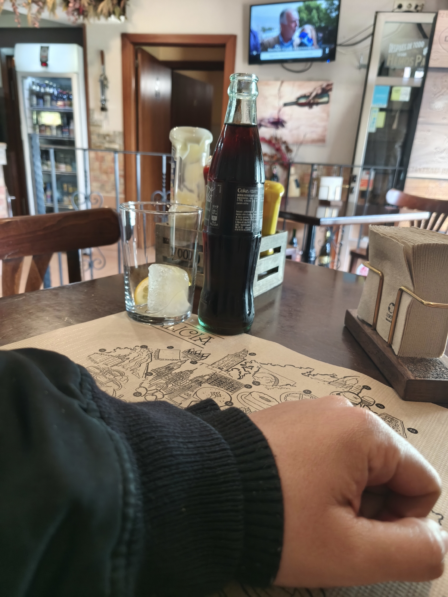 The image portrays an indoor scene, likely within a casual restaurant, bar, or cafe in Pontevedra, Spain. In the foreground, a person's left forearm and hand, clad in a dark, ribbed sleeve, rest on a brown paper placemat. The placemat features a detailed black-and-white illustration resembling a city map or architectural landmarks, with visible text including "KLICIA" and several numbers like "18", "47", "1000", and "1001".
On the dark wooden table, a classic glass bottle of Coca-Cola stands, its label displaying "Coke" and Spanish text "BEBIDA REFRESCANTE AROMA y aroma cafeína." Adjacent to it is an empty drinking glass containing ice cubes and a lemon slice, ready for the beverage. A small wooden caddy holds two condiment bottles, one yellow and one white, while a metal and wooden napkin holder stacked with brown napkins is positioned to the right.
The background reveals a bustling interior. To the left, a tall white refrigerator or cooler filled with various bottles and items is visible. Above the table, a flat-screen television mounted on the wall displays a program featuring two individuals, an older man and a woman. Wall decorations include a painting depicting wine being poured and a narrow vertical display case with Spanish text "DESPUES DE TODO" and "HUHOS PA" (likely "HECHOS PARA"). A wooden doorway and chairs are also part of the rustic-themed setting, complemented by dried foliage hanging from above. The scene suggests a relaxed moment, possibly awaiting or enjoying a drink.