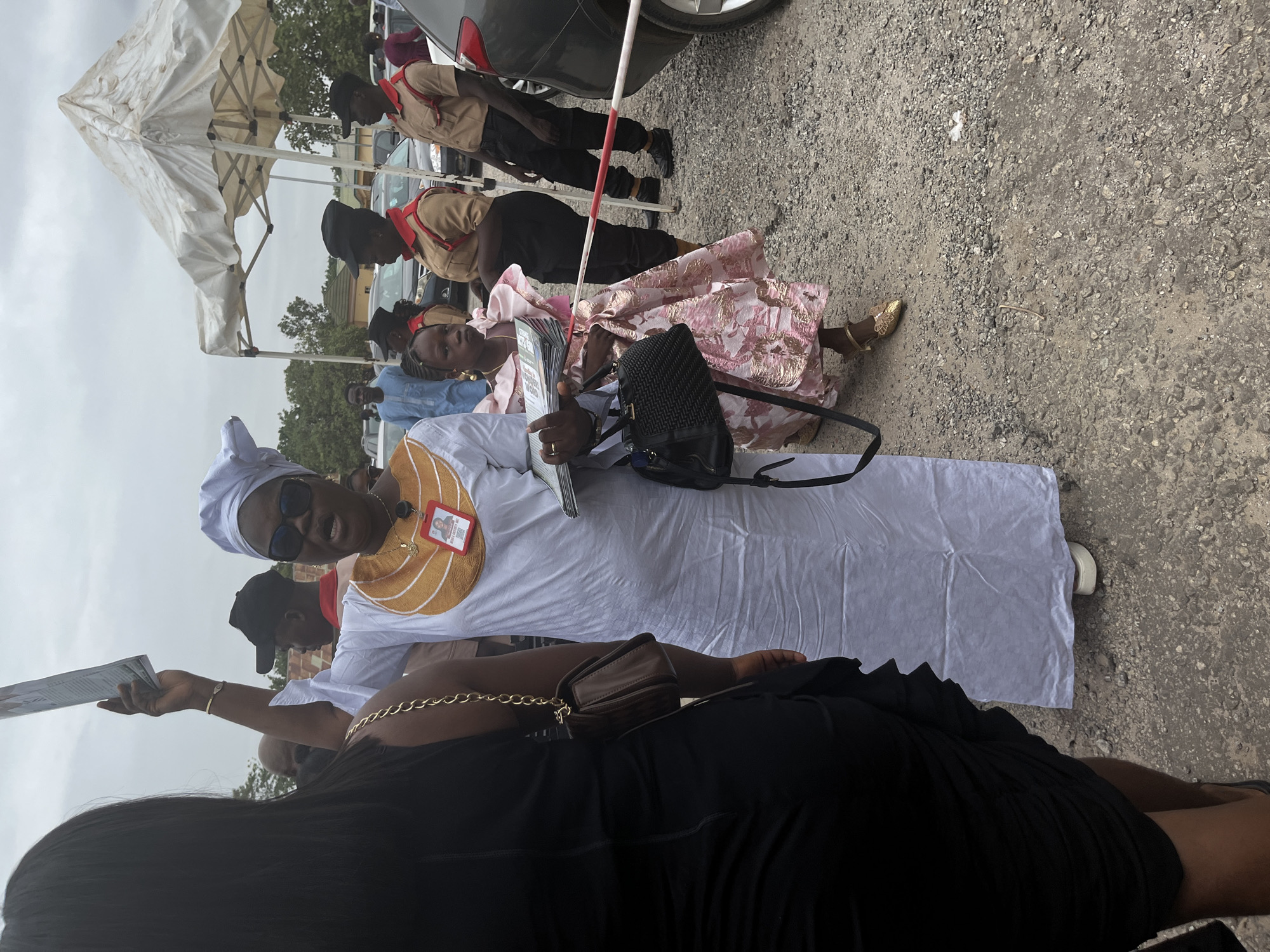 The image depicts a gathering outdoors in Abuja, Nigeria, on what appears to be a cloudy day. A woman in a white, floor-length dress and a matching headwrap is holding a pamphlet or printed material in her hand. She is wearing sunglasses and has a name tag pinned to her dress. Another woman in a pink floral dress is kneeling nearby, also holding printed material. Behind them, a group of individuals in scout uniforms can be seen standing in a line. A white tent is visible in the background, along with a few cars parked nearby on a paved surface.