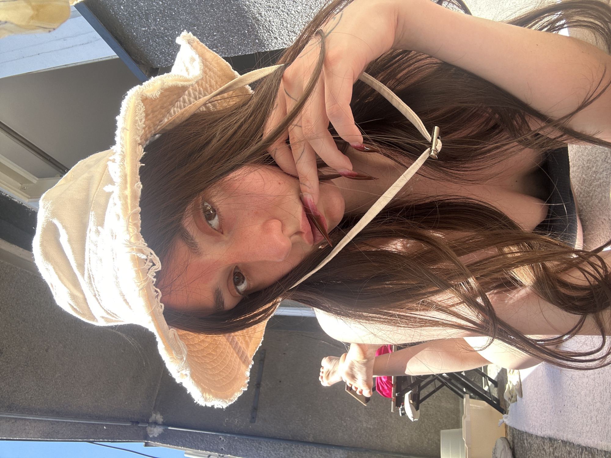 A woman wearing a beige sun hat is seen in an outdoor setting. She has long, brown hair and is holding her hand up near her face, with long, manicured fingernails visible. Her body is reclined, suggesting she may be sitting or lying down. Visible in the background are parts of the outdoor environment, including what looks like a concrete or textured wall, suggesting a roof terrace, yard or balcony. The angle of the shot is from above, pointing downwards. The scene is located in Vracar Urban Municipality, Serbia. The lighting is bright, indicating a sunny day.