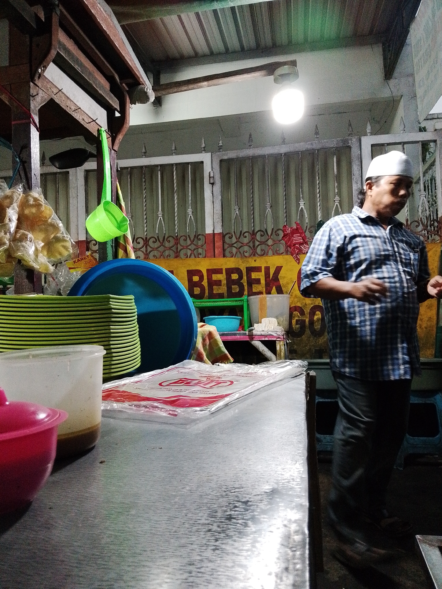 The image depicts an outdoor food stall or eatery, likely in Pasuruan, Indonesia, during nighttime or low light conditions. In the background, a man wearing a white peci (a brimless cap) and a blue checkered shirt is visible. He appears to be engaged in a transaction or serving customers, with his hands in motion. Behind him, a yellow sign with red lettering reads "BEBEK GORENG," indicating that duck is the primary dish served. The stall features various food-related items, including stacks of green plates, a large blue plastic bowl, and bags of snacks or chips hanging to the left. A bright, bare light bulb illuminates the area, casting shadows. The foreground shows a metallic countertop with a pink bowl, a white container, and what appears to be plastic packaging for food items. The overall impression is of a busy, informal dining establishment.