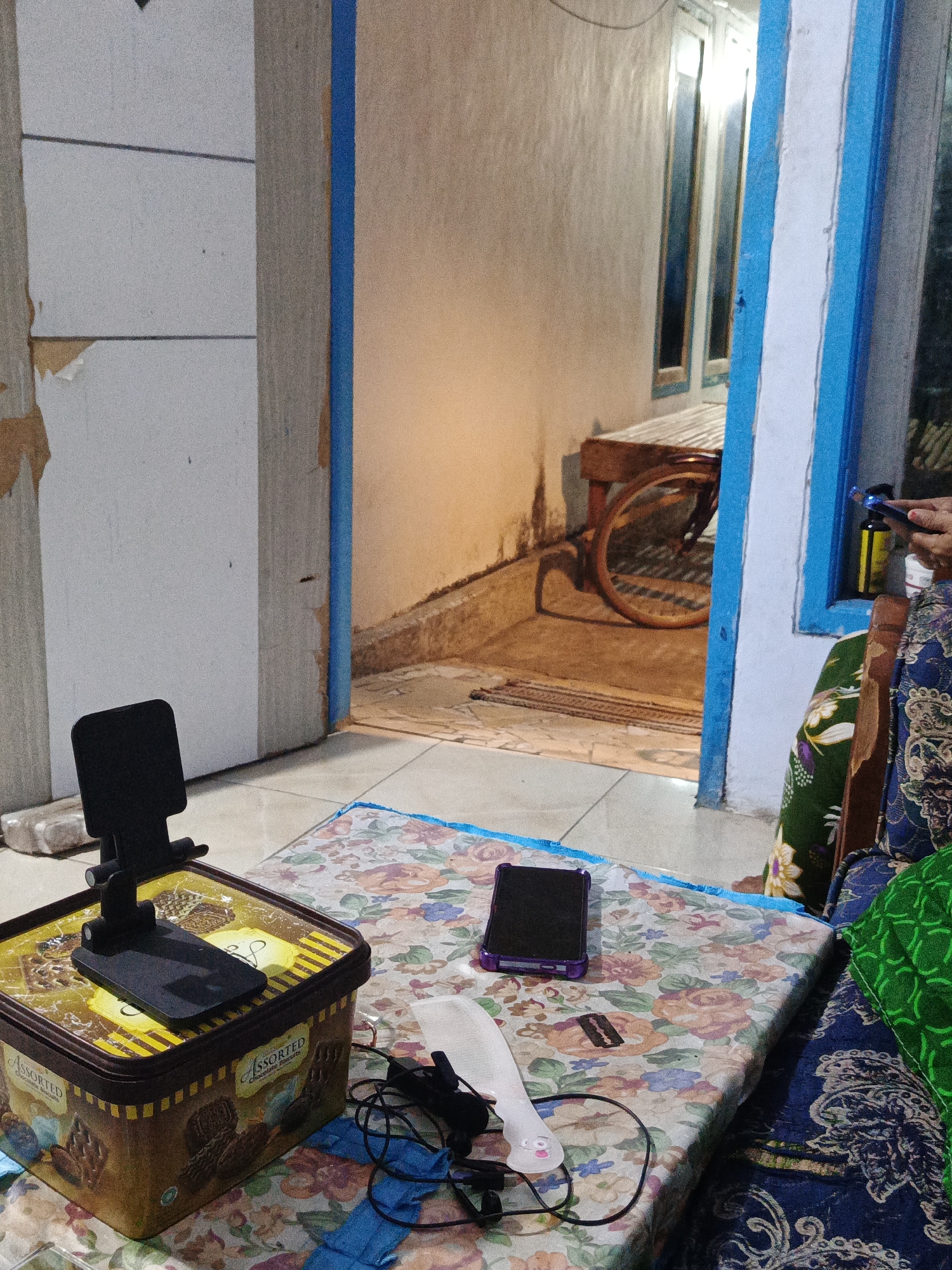 The image depicts an indoor scene with an open doorway leading to an outdoor area. In the foreground, a patterned cloth covers a surface, on which rests a black smartphone stand holding a black smartphone, a black smartphone with a purple case, charging cables, and a white object with a pink smiley face. To the left, a brown tin container with "Assorted Chocolate Biscuits" printed on it serves as a base for the smartphone stand. The walls of the indoor area are a light color, with some peeling wallpaper. The doorway reveals an outdoor area with a woven wooden bench and a bicycle wheel visible. A person is partially visible on the right side of the image, wearing a blue and patterned garment, and holding a yellow bottle. The overall impression is a casual domestic setting, possibly in the city of Sumbersari, Indonesia, given the cultural context suggested by the decor and furnishings.