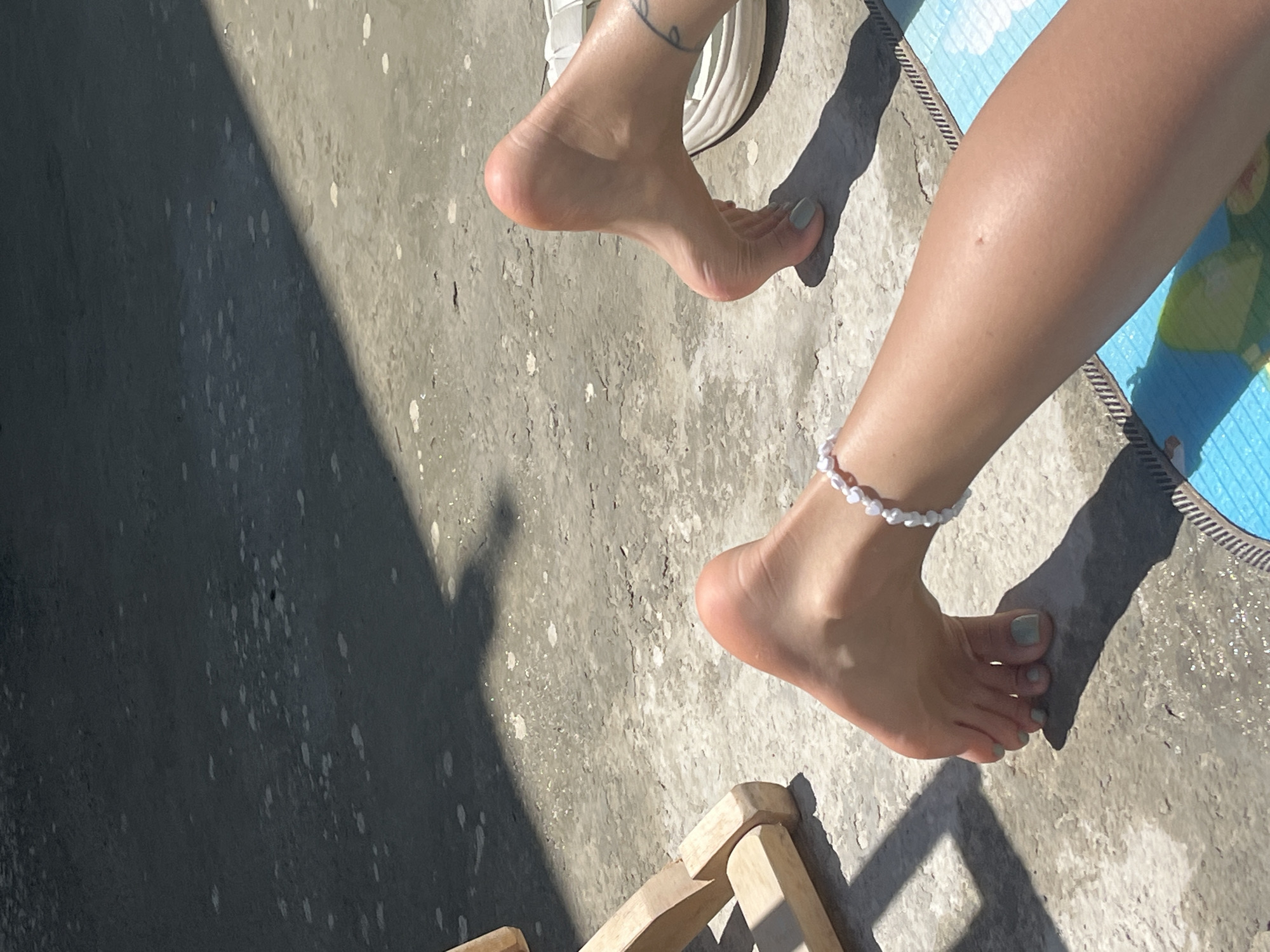 The image displays two bare feet on a concrete surface. The left foot, closer to the viewer, has a pearl anklet on it and is positioned with the toes pointed downwards. The right foot is further away and angled slightly. A shadow from an unseen object falls across the concrete. In the background, a portion of a wooden chair is visible to the left. The scene appears to be outdoors on a sunny day.