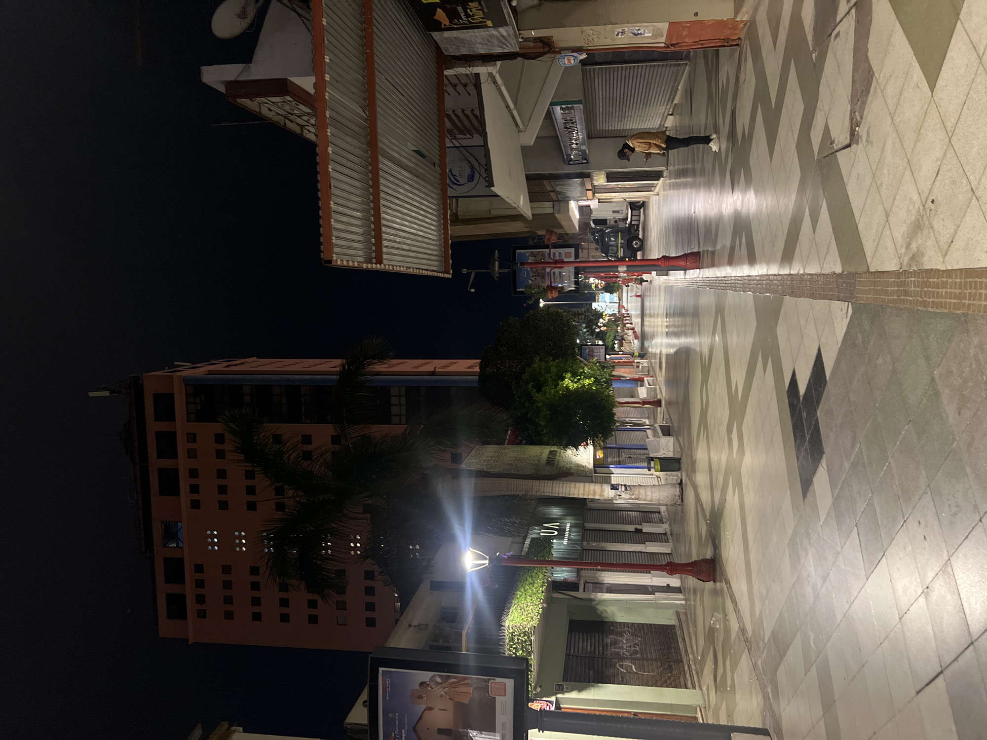 Arica, Chile. A street scene at night. A high-rise building, mostly orange, is on the left side of the image. The view is along a pedestrian promenade. On the right and left of the promenade, shops are closed. A person in a jacket and dark headwear walks near the edge of the promenade. Red, decorative lampposts line the walkway. A large sign is in the lower left corner.