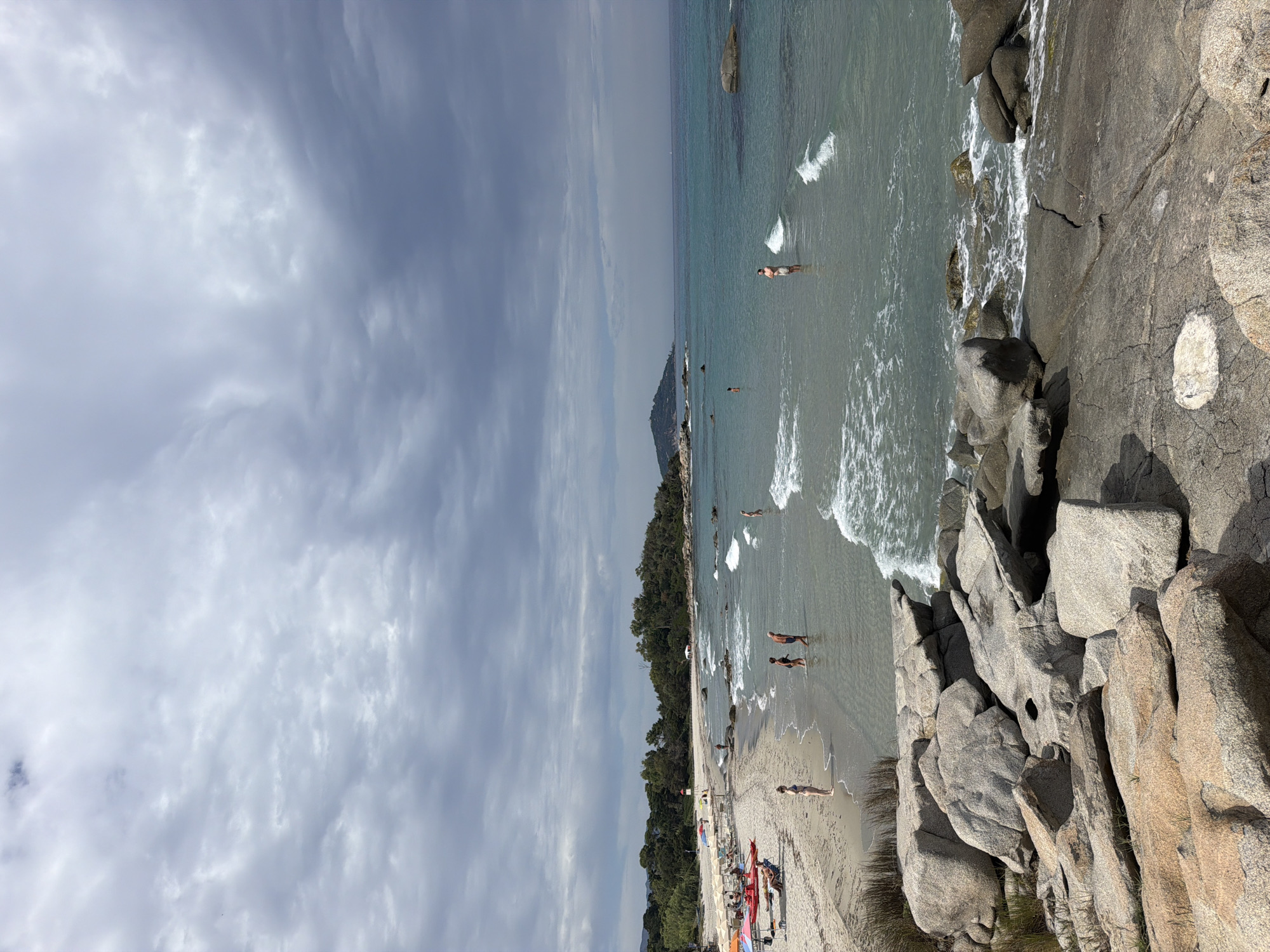 The image depicts a scenic view of a beach in Tortolì, Italy. The foreground is dominated by large, weathered granite rocks. The middle ground shows a sandy beach with several beach umbrellas and lounge chairs, some with people relaxing or seated. A line of people are visible walking along the shoreline or wading in the shallow, clear turquoise water. Further out in the sea, more people are present, some standing in the water and one walking towards the shore. The background features a densely wooded coastline with trees and vegetation, leading to a hilly landscape. The sky is overcast with heavy, grey clouds, suggesting a potentially stormy or cloudy day. The overall scene captures a tranquil beach environment with a mix of natural rocky formations and human activity.