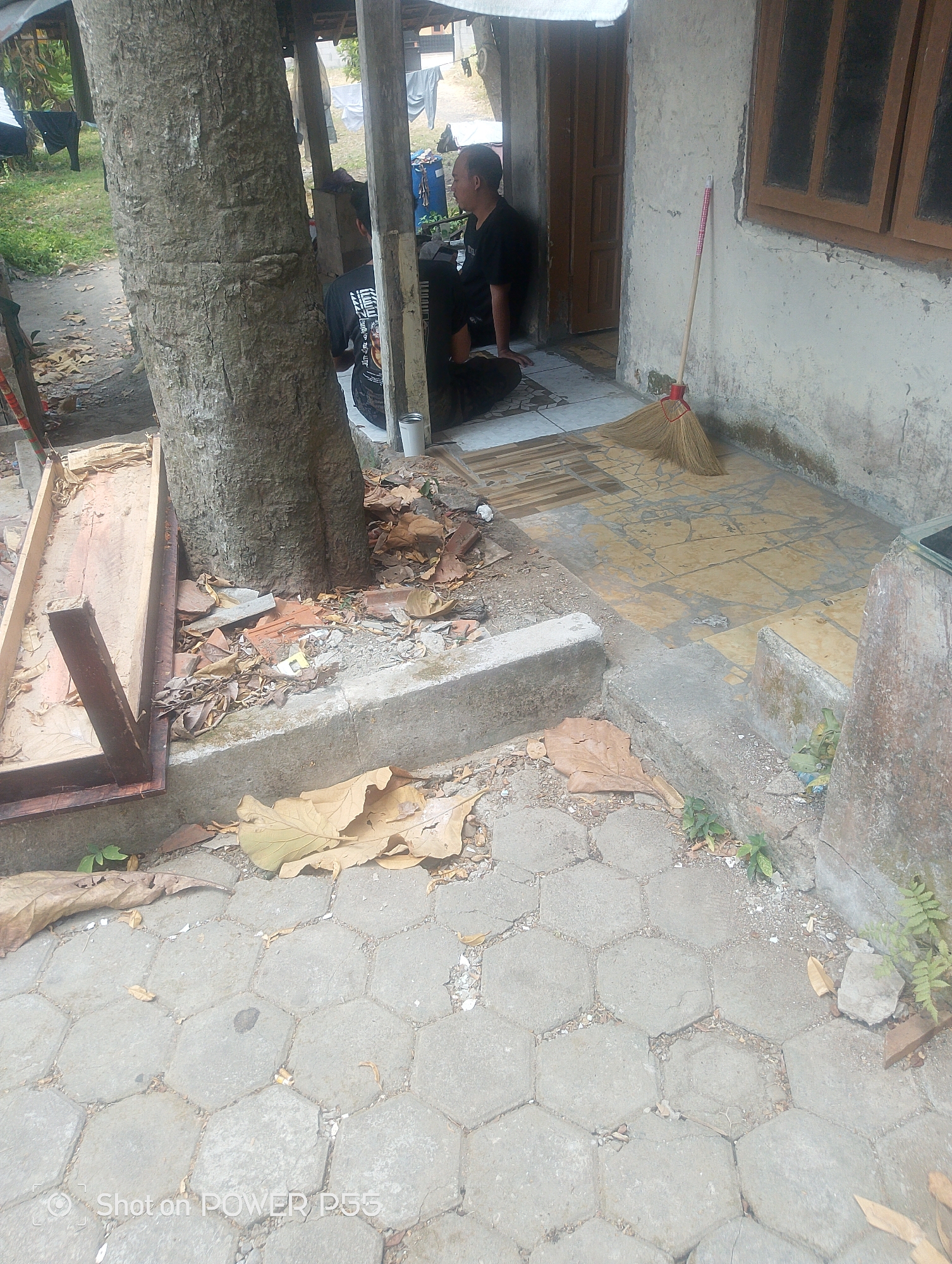 The image depicts an outdoor scene in Lebaksiu, Indonesia, focusing on a pathway leading to a building with a textured, light-colored wall and a wooden-framed window. A large tree trunk stands prominently on the left side of the frame, with a wooden structure resembling a dismantled table or bench situated beside it. The ground is a mix of hexagonal concrete pavers and natural earth, littered with dry leaves and some debris, including broken pottery or tiles.
In the background, a person is seated on the ground in front of the building, facing away from the camera and towards the left. This individual is wearing a dark t-shirt with a graphic print. Another person is partially visible to the right of the seated individual, also appearing to be seated, wearing a dark shirt. Laundry hangs on a line in the far background, suggesting a residential area.
In front of the building's wall, a broom with a red handle and tan bristles rests on a tiled section of the pathway. The tiled area transitions from the hexagonal pavers. The overall environment suggests an unkempt outdoor space adjacent to a simple dwelling.