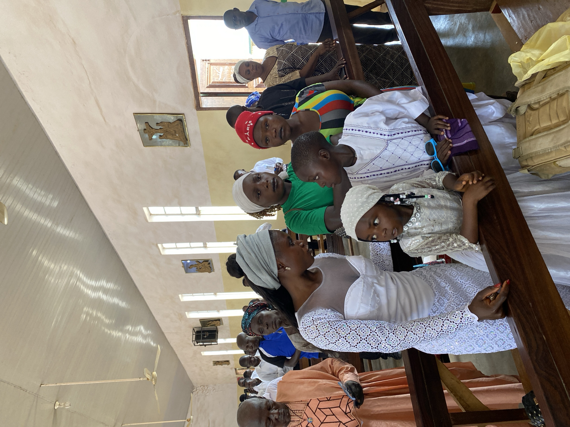 In Kuru Dungus, Nigeria, an indoor scene depicts a group of people sitting on wooden benches inside a building, possibly a church or religious gathering place. The room is well-lit by several windows along the walls. A woman with a gray head covering is seated prominently near the center, wearing a white dress with a lace-like pattern on the sleeves. She is looking toward another person. To her right, a child wearing a white head covering and dress looks upward. To the left and further back, other people are visible, some wearing brightly colored clothing, including a red hat. A wooden structure or pew divider is visible on the right side of the image. A bag or other object rests on the bench. A fan, windows, and religious decorations are visible on the ceiling and walls.