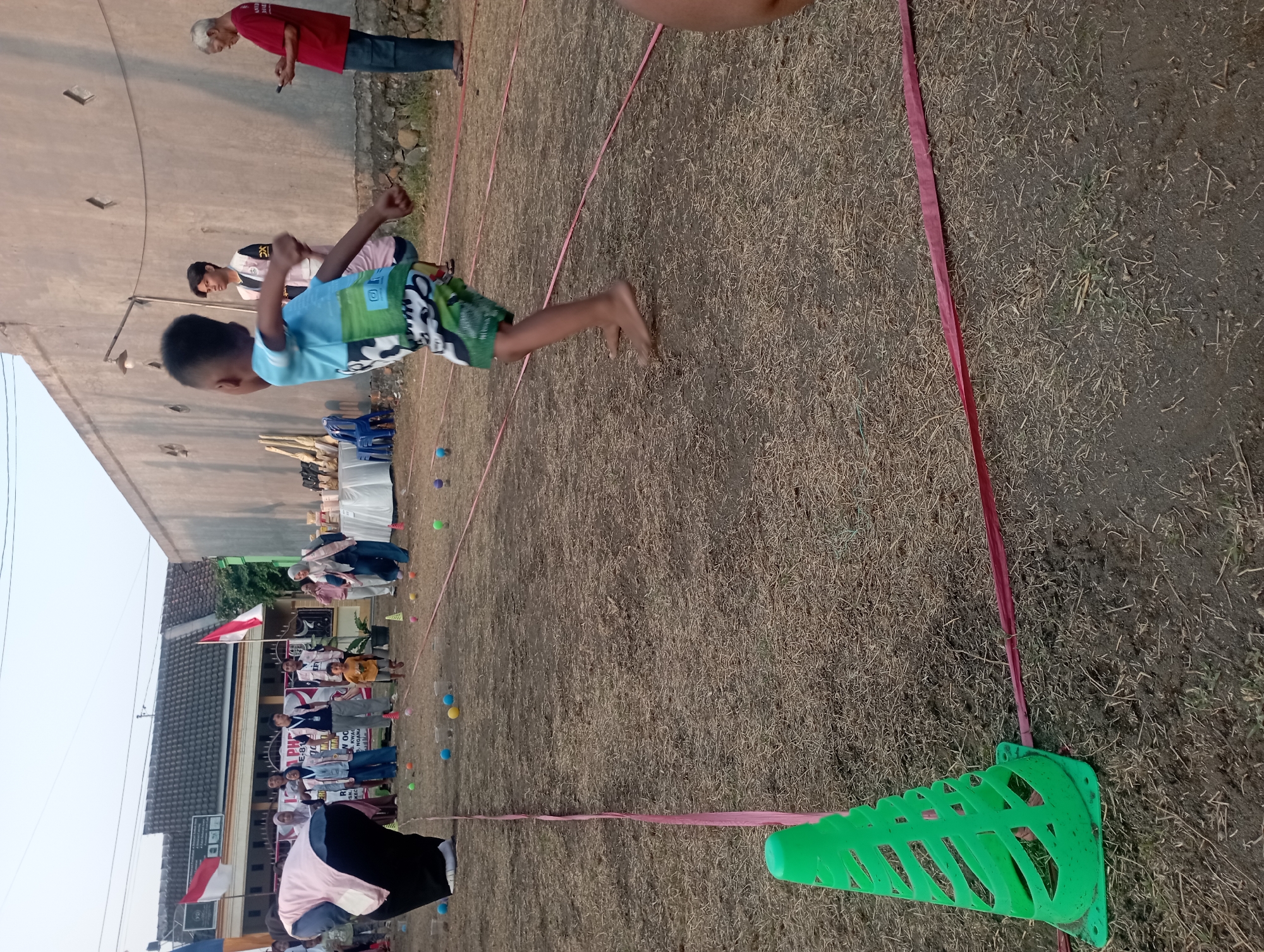 The image depicts an outdoor event, possibly a children's competition or festival, taking place on a dry, grassy field in Candirejo, Indonesia. A young boy, the primary subject in the foreground, is captured mid-action, appearing to throw or jump with his right arm extended. He is wearing a blue and green patterned t-shirt and green shorts, and is barefoot.
Several other individuals are visible in the background, including adults and children. Some adults are wearing vests, suggesting they are organizers or participants in some capacity. There are also Indonesian flags displayed, and a table with trophies or prizes set up. The field is marked with pink ribbons or tape, creating lanes or boundaries for the event. A bright green cone stands on the left side of the frame. The environment suggests a warm, possibly late afternoon setting, with a building and a concrete wall forming the backdrop.