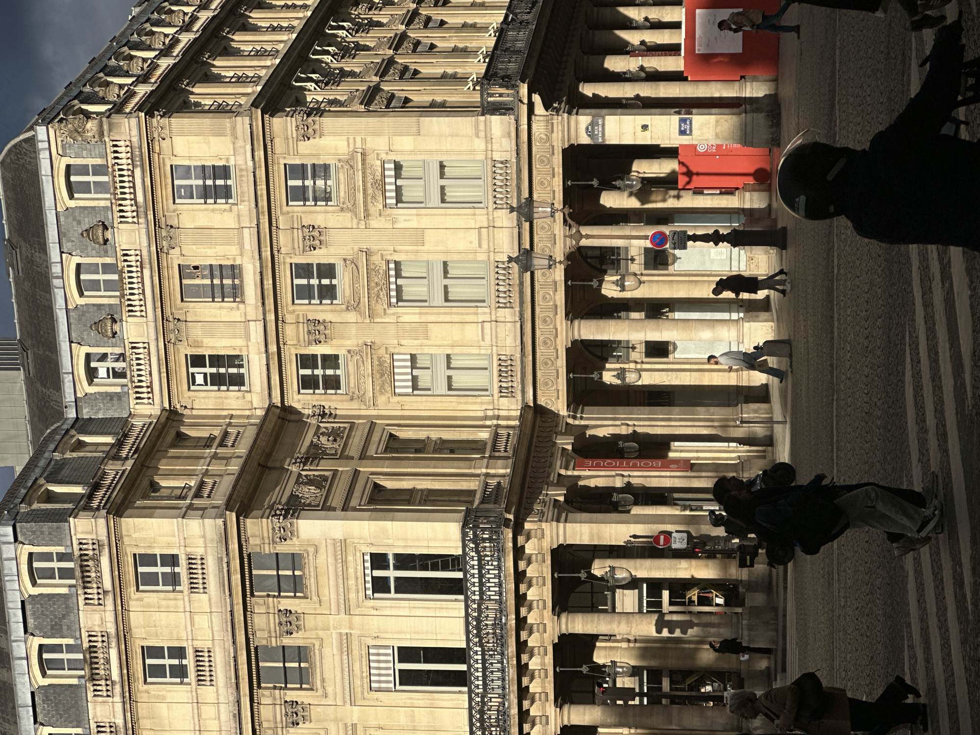 The image depicts a street scene in Paris, France. A tall, ornate building with multiple windows and architectural details dominates the left side of the frame. The building's facade is light-colored, and there's a balcony visible on one of the floors. On the right, a colonnade with evenly spaced columns runs along the street. People are walking on the sidewalk in front of the building and colonnade. Various street furniture, including streetlights and signposts, are visible. The image appears to have been taken in the afternoon.