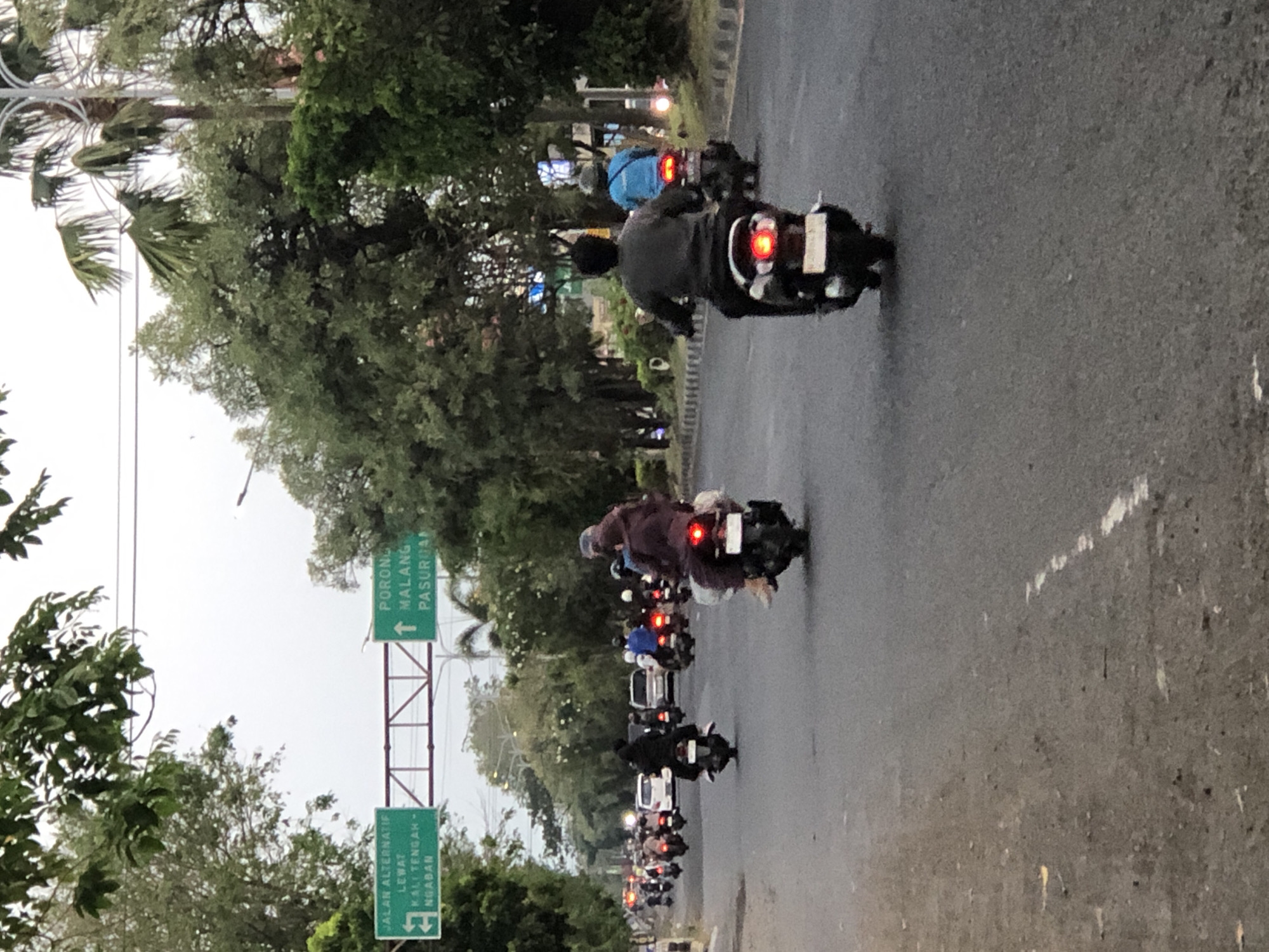 The image depicts a street scene in the city of Gelam, Indonesia. Several individuals are riding motorcycles on a paved road. The road appears to be a main thoroughfare, with trees lining the sides and overhead traffic signs. The sky is overcast, suggesting it may be early morning or late afternoon, or a cloudy day.
On the left side of the road, a green traffic sign indicates "JALAN ALTERNATIF" (Alternative Road) with directions to "LEWAT KALI TENGAH - NGABAN" (Pass Kali Tengah - Ngaban). On the right side of the road, another green traffic sign points upwards and reads "PORONG MALANG PASURUAN" (Porong Malang Pasuruan), indicating directions to these cities.
In the foreground, the back of a motorcycle rider is visible, wearing a dark jacket and a helmet. Behind them, another motorcycle rider in a blue jacket is also seen. Further down the road, multiple other motorcycles are present, some with passengers. There are also a few cars visible in the distance. The overall impression is a busy urban street with moderate traffic.