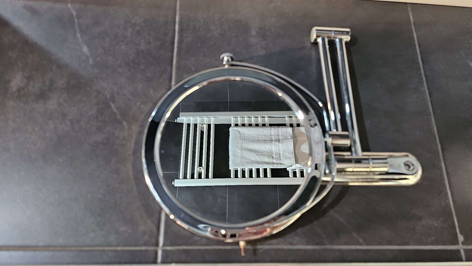 This is a close-up shot of a chrome magnifying mirror mounted on a dark gray tiled wall. The mirror reflects a white towel rack with a light gray towel hanging from one of its rungs. The mirror is round with a double-jointed arm extending from its side, attached to a mounting bracket on the wall. The background consists of dark gray tiles with visible grout lines. The lighting suggests an indoor setting, likely a bathroom. No people or specific locations in Alhama de Aragón, Spain are identifiable in the image.