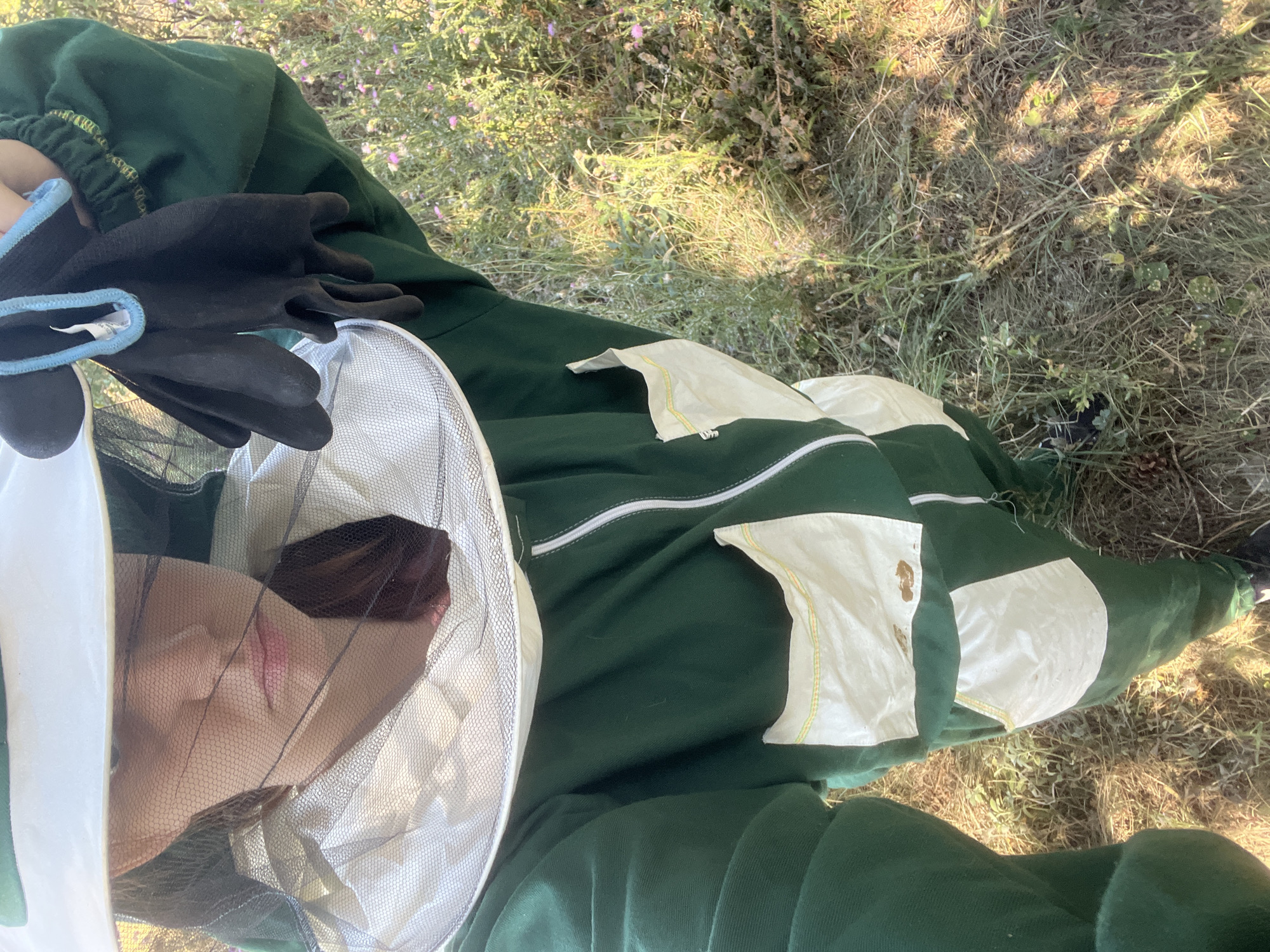 A person, clad in a full green beekeeping suit, stands outdoors in a grassy area. The suit features a white veil with mesh netting that obscures the wearer's face, a wide-brimmed hat, and black beekeeping gloves held in the right hand. The suit has several cream-colored pockets with yellow trim. The background consists of dry grass and small wildflowers, bathed in sunlight. The image appears to be taken at ground level, looking up at the beekeeper. This scene is located in the city of Davidovo, Bulgaria.