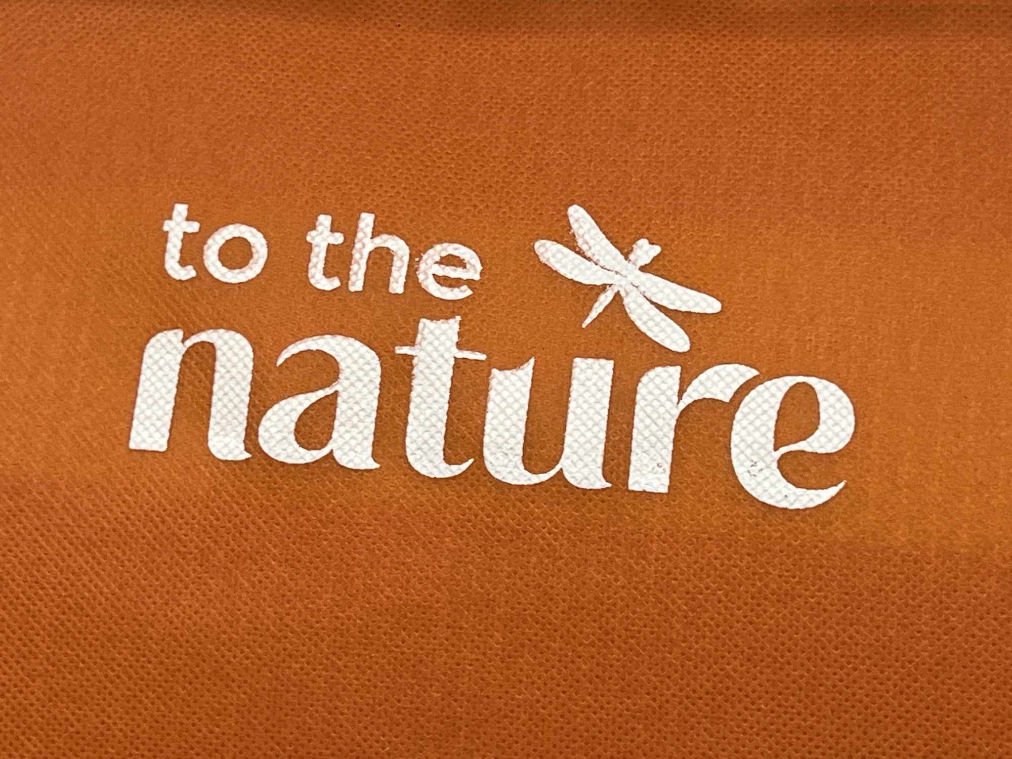 The image is a close-up shot of white text and a white dragonfly graphic printed on an orange textured surface, likely fabric. The text reads "to the nature". The dragonfly is positioned to the right of the word "the" and above the word "nature". The orange surface has a visible woven pattern. The image does not provide any information about the location of this item in the city of Ulyanovsk, Russia.