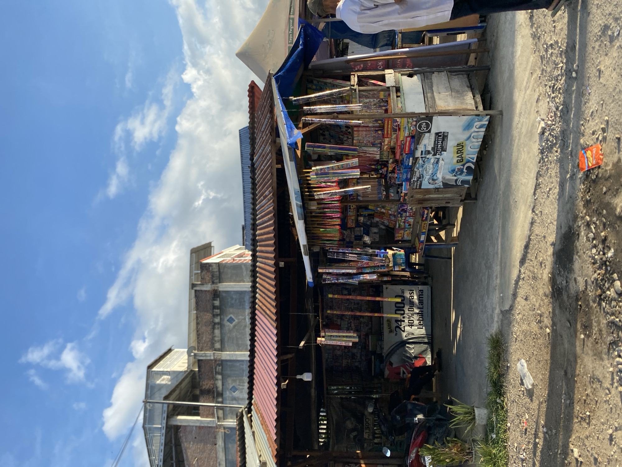 The image shows a roadside stall in Lhokseumawe, Indonesia. The stall is constructed with a corrugated metal roof and wooden supports. It is stocked with various goods, including fireworks and cigarettes (brands such as Dunhill and Sampoerna A Mild, referencing their 24.000 variant). Signage promotes a cigarette offering "40% Durasi Lebih Lama" (40% Longer Duration). A parked red motorcycle is visible in front of the stall. The stall is on a paved surface next to a dirt and gravel shoulder. In the background are buildings, and the sky is partly cloudy. A person is partially visible standing near the front of the stall.