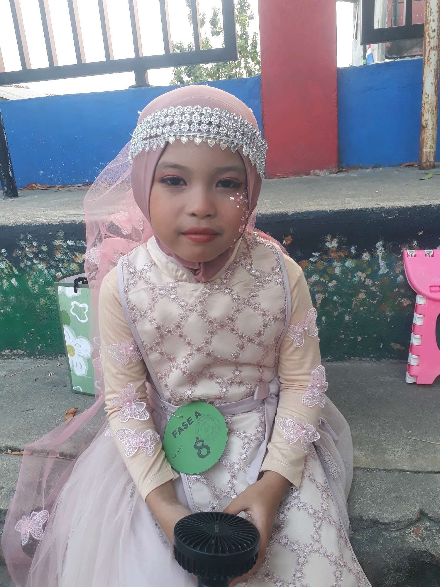 A young girl, adorned in a light pink hijab and a matching ornate dress with butterfly accents, sits on a concrete ledge outdoors. Her hijab is embellished with a sparkling silver band. Her face, painted with subtle makeup including glitter accents near her eyes, looks directly at the camera. She holds a small black fan in her hands. A green circular tag with the text "FASE A" and the number "8" is pinned to her dress. Behind her, a blue and red wall forms the backdrop, with a metal fence visible above. A green and white patterned bag rests to her left, and a pink plastic stool is to her right. The ground is a rough, grey concrete surface. The scene appears to be in an outdoor public space within the city of Pangkalpinang, Indonesia.