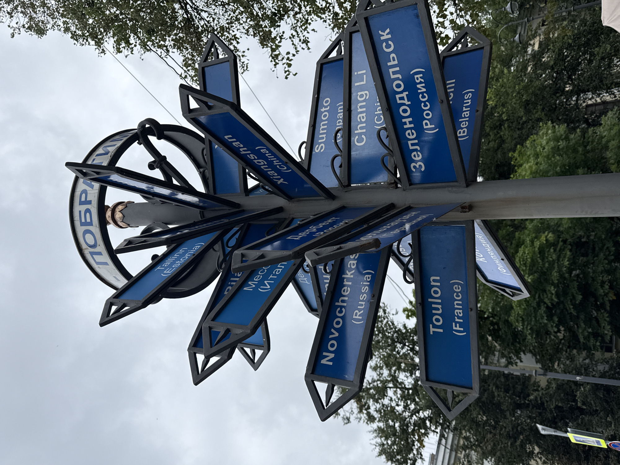 A low-angle medium shot shows a directional signpost in Kronstadt, Russia. The signpost is made of metal and has multiple blue directional signs pointing in various directions. The signs list city names and their respective countries, some in English and some in Russian. Examples of destinations include "Tallinn (Estonia)", "Mecca (Italy)", "Novocherkassk (Russia)", "Xiangshan (China)", "Sumoto (Japan)", "Chang Li (China)", "Zelenodolsk (Russia)", "Toulon (France)", and "Belarus". At the top of the signpost, a circular sign with blue lettering reads "ПОБРАТИМ" which translates to "Sister City". The sky is overcast, and trees are visible in the background. The signpost is the primary subject, with the surrounding environment providing context. No people are visible in the image.