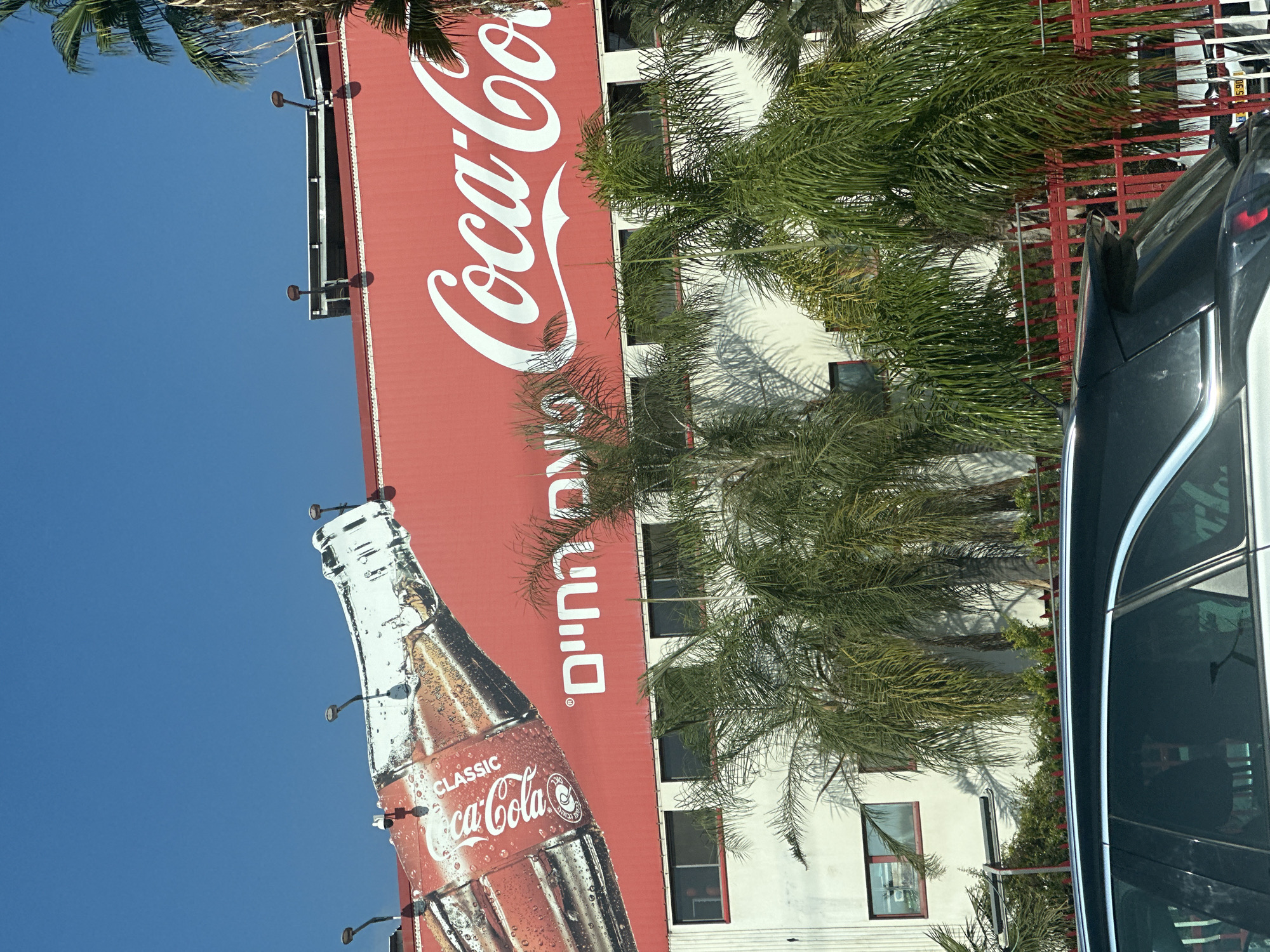 The image shows a large Coca-Cola advertisement on the side of a white building. The advertisement features a giant Coca-Cola bottle and the Coca-Cola logo in white script on a red background. To the left of the Coca-Cola logo, there is writing in Hebrew. The building appears to be an industrial or commercial facility, with several windows visible. Palm trees are in the foreground, obscuring parts of the building and advertisement. The sky is clear and blue. In the very bottom of the image, the rear of a dark-colored car is visible, with a red fence behind it. This scene is located in the city of Bnei Brak, Israel.