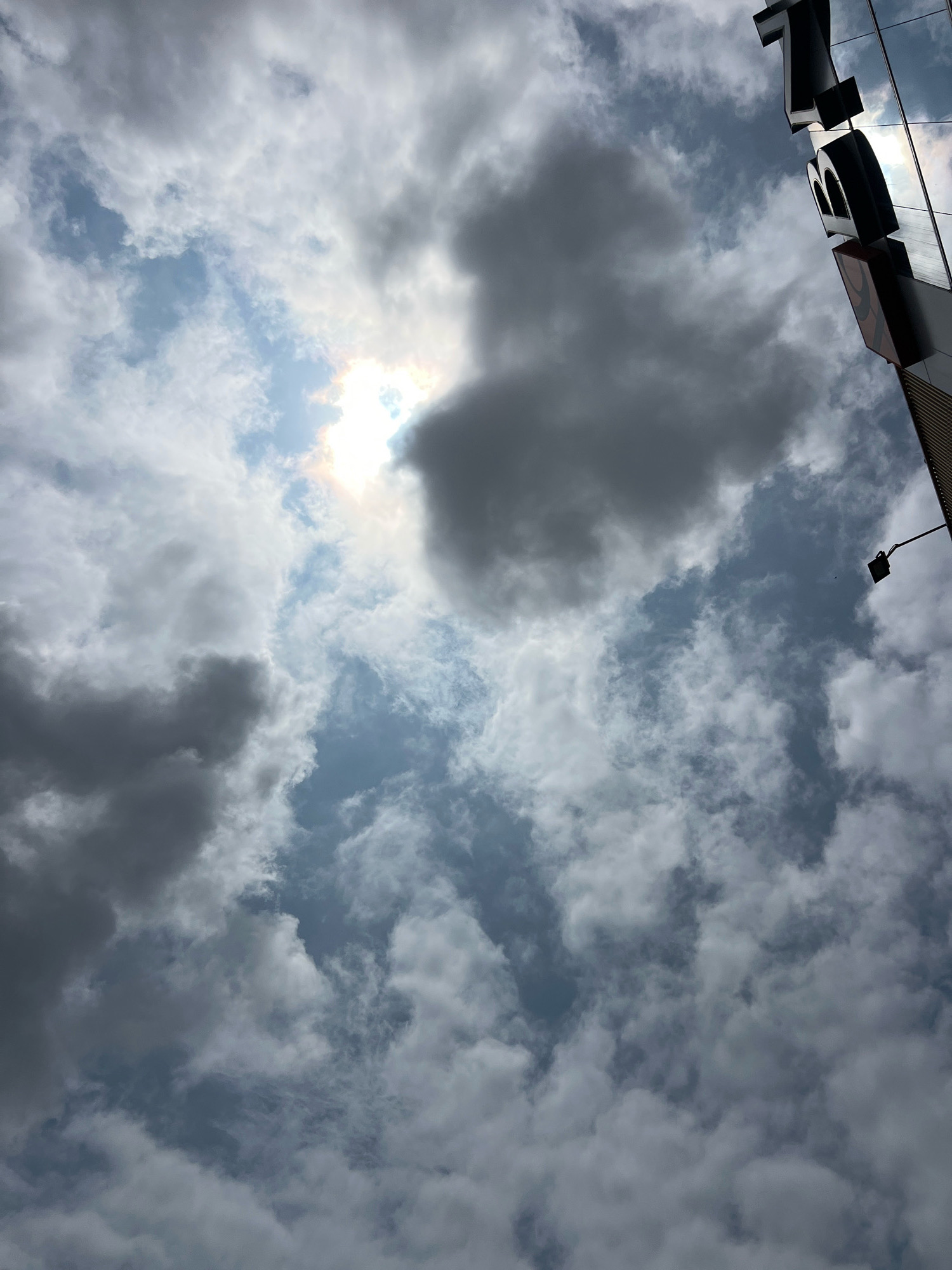 The image captures a sky filled with dramatic clouds, with the sun partially obscured by them, creating a bright, diffused light. In the upper right portion of the frame, a portion of a building is visible, featuring large, three-dimensional black letters that appear to spell out "ZION." The building's facade is reflective, mirroring the cloudy sky. A street lamp with a dark rectangular fixture is also visible mounted on a pole, extending from the right side of the frame. The overall scene suggests an outdoor urban environment with a dynamic sky. This image was taken in Denpasar, Indonesia.