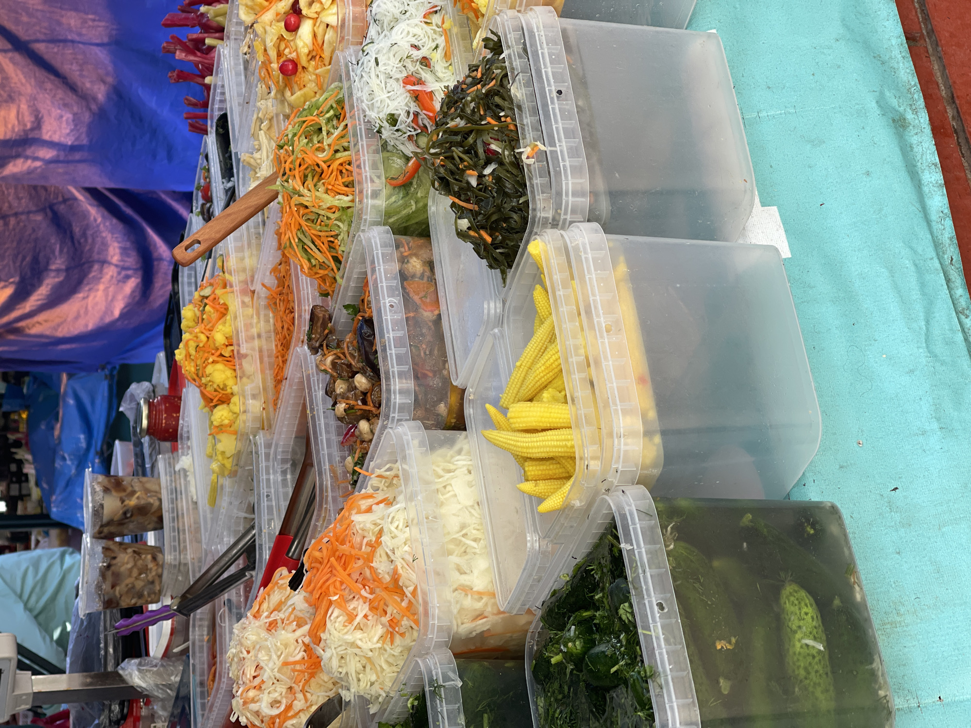 The image shows a display of food items in clear plastic containers arranged in rows, likely at a market. The items include shredded carrots and cabbage, baby corn, pickled cucumbers, and various prepared salads. A wooden spoon and tongs are visible, suggesting customers can serve themselves. The background includes a blue tarp and glimpses of other market stalls. The image is taken at an angle. The location and time period are Unknown.