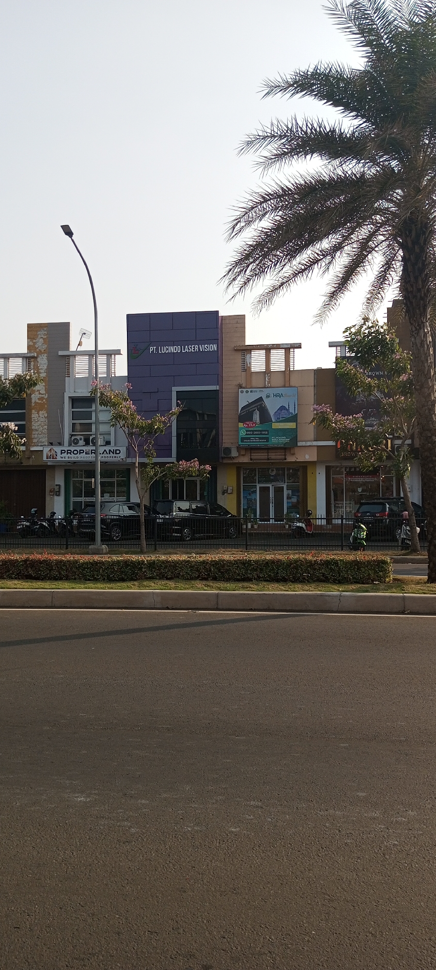 The image captures a street scene in Lambangjaya, Indonesia, featuring a row of modern commercial buildings. The buildings are of varying heights and architectural styles, with prominent signage indicating businesses. One sign clearly displays "PT. LUCINDO LASER VISION." Another visible sign reads "PROPER.LAND WE BUILD PROPERTY PROPERLY." The buildings are fronted by a paved sidewalk and a low hedge, beyond which is a road. Several vehicles, including cars and motorcycles, are parked or in motion on the street. A tall palm tree with distinctive fronds dominates the right side of the frame, partially obscuring the sky. The sky is clear and light, suggesting daytime. A street lamp with a curved arm is visible on the left side of the image. The overall impression is one of an active urban commercial area.