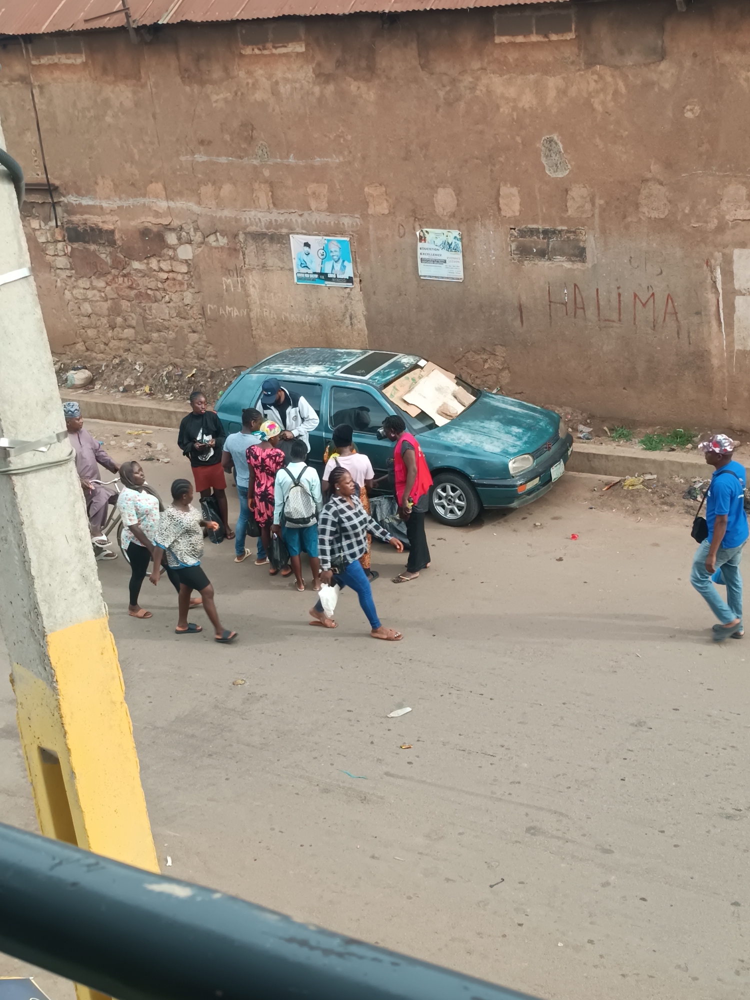 The image, taken at an elevated vantage point, depicts a street scene in Bukuru, Nigeria. A group of approximately 14 people of various ages and attire are either walking across or standing on the paved road. A dark teal Volkswagen Golf, bearing damage including cardboard in place of a windshield, is parked along the roadside in front of a tall, weathered wall. The wall has visible patches of repair, some bare brick, and faded graffiti, including the word "HALIMA". Two posters are affixed to the wall, above the vehicle. One man walks alone on the right side of the frame, wearing a blue shirt and jeans. On the left, a yellow and gray post or column, part of a structure like a balcony or bridge, is visible in the foreground. The overall impression is of a bustling, somewhat congested urban street.