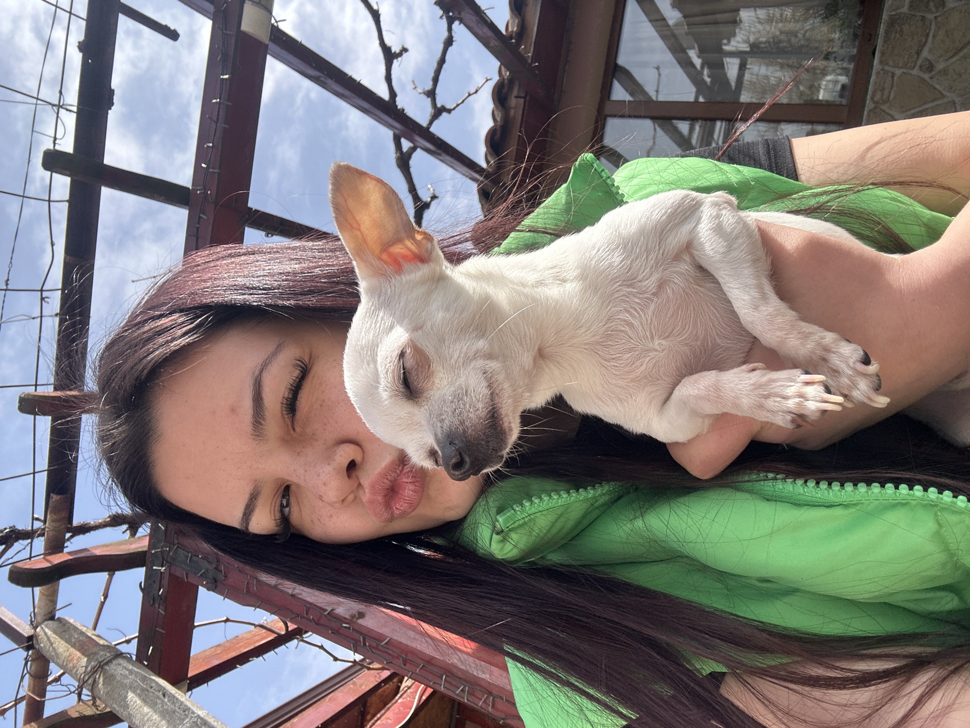 A woman is holding a small, white dog. The woman has long, dark hair, closed eyes, freckles, and is making a kissing face. She is wearing a bright green jacket. The dog appears to be sleeping. A metal structure, possibly a trellis or a pergola, is visible in the background, along with a partially visible building and a blue, cloudy sky. The location is Chubra, Bulgaria.