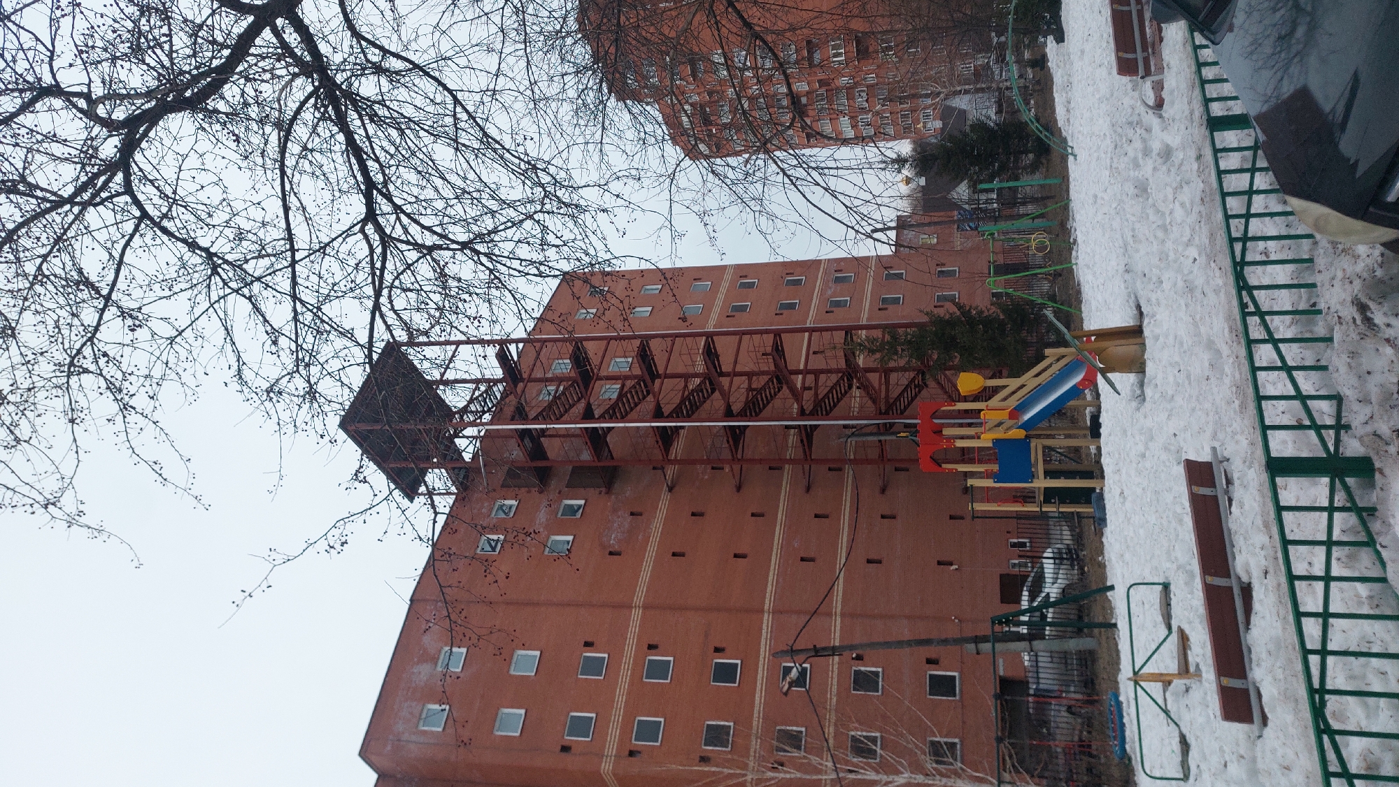 The image shows a residential area in Novosibirsk, Russia, likely during the late winter or early spring. An orange-brick apartment building dominates the frame, featuring rows of square windows and a large, metal fire escape structure extending along its side. The playground located at the foot of the building, covered in snow and includes a slide, climbing structures, and benches. Bare branches from deciduous trees reach into the frame from above and to the left. Another apartment building is visible in the upper right corner. The sky is overcast, suggesting a cold or gloomy day.