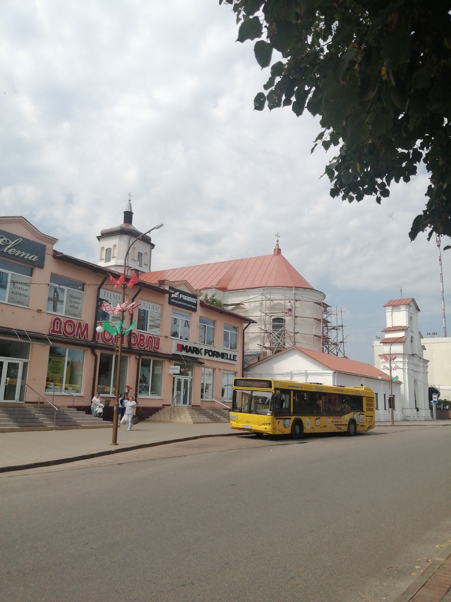 The image depicts a street scene in Slonim, Belarus, featuring a yellow bus, a pink building with shops, and church structures. The bus, a model likely used for public transportation, is prominent in the foreground, traveling on a paved road. The pink building, situated on the left side of the image, displays several shop signs in Cyrillic, including "Elena," "Дом торговли" (House of Trade), and "Mark Formelle." One of the shops appears to be under renovation or construction, with scaffolding visible on its exterior.
Several people are visible on the sidewalk in front of the pink building, some walking and others standing. They are dressed in casual attire. Behind the pink building, a church with a red roof and white walls is partially visible. Further to the right, another white church structure with a steeple and red roof is also present. The sky is overcast with clouds. The overall environment suggests a typical urban setting in a town or city. The presence of shops and a bus indicates commercial activity and public infrastructure.