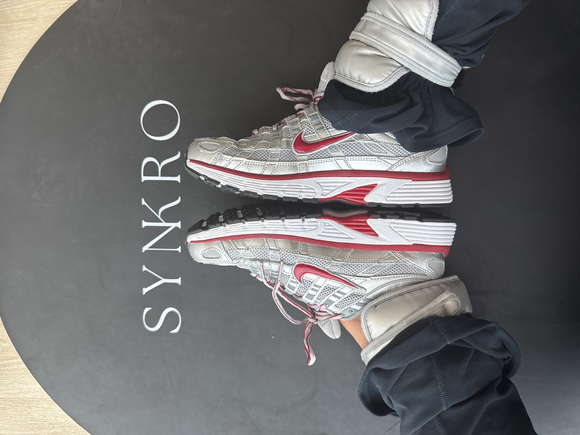 This is a top-down, eye-level, full shot of a person's feet wearing silver and red Nike sneakers. The sneakers are on a black circular mat with white lettering that reads "SYN KRO". The person is wearing black joggers. The image does not contain any information about the location in Dubai, United Arab Emirates.
