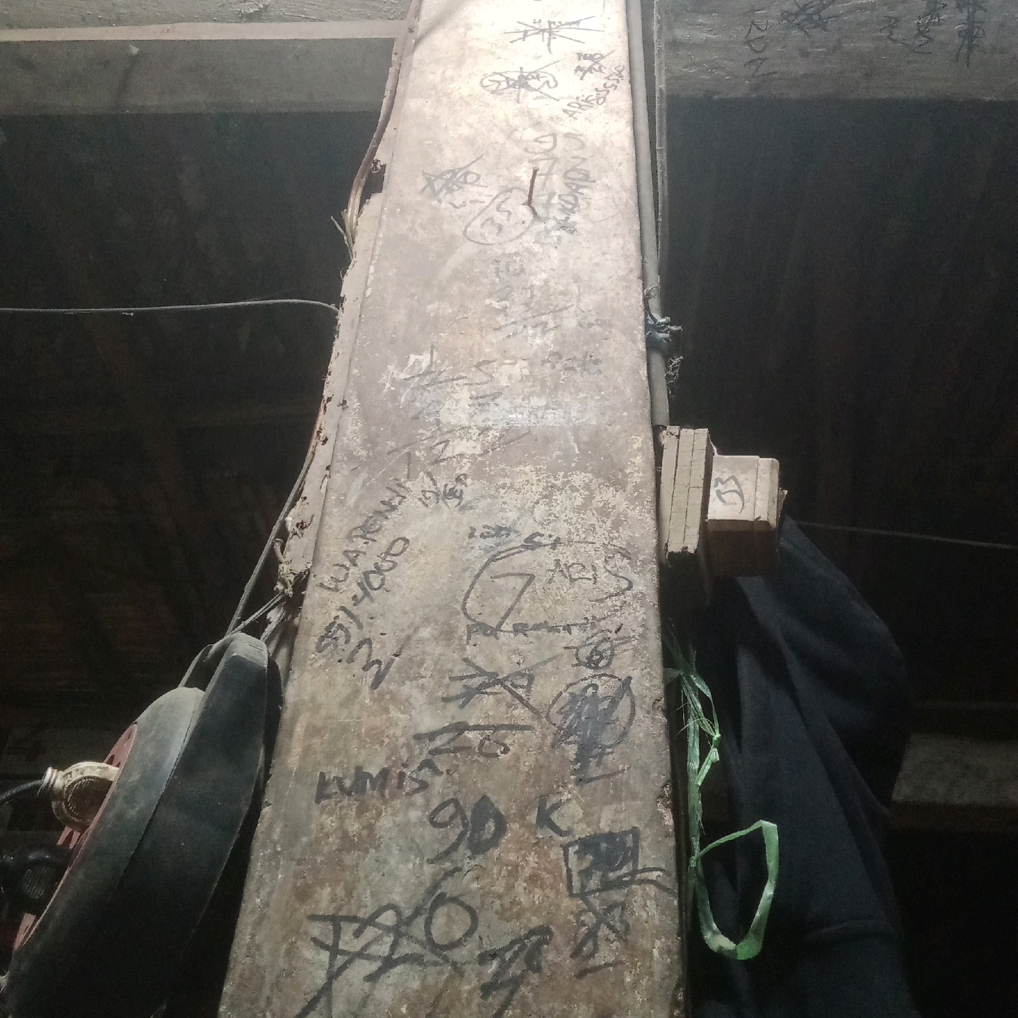 A vertical, weathered metal beam, possibly a support column, is heavily covered in graffiti. The graffiti consists of various tags, numbers, and stylized drawings in black and gray markings. Some of the markings are difficult to decipher due to the surface texture and the overlapping nature of the graffiti. Towards the bottom left of the image, a large, dark, circular object, possibly a tire or a part of machinery, is leaning against the beam. To the right of the beam, a dark fabric, possibly clothing or a tarp, is draped. Near the top of the beam, a small wooden block with the number "33" written on it is attached. The background appears to be a dimly lit, industrial or storage space, with visible structural elements like wooden beams and dark walls. The overall impression is of an urban, possibly abandoned or neglected area, consistent with a location in a city like Karawang, Indonesia, where such graffiti is common in industrial zones or underpasses.