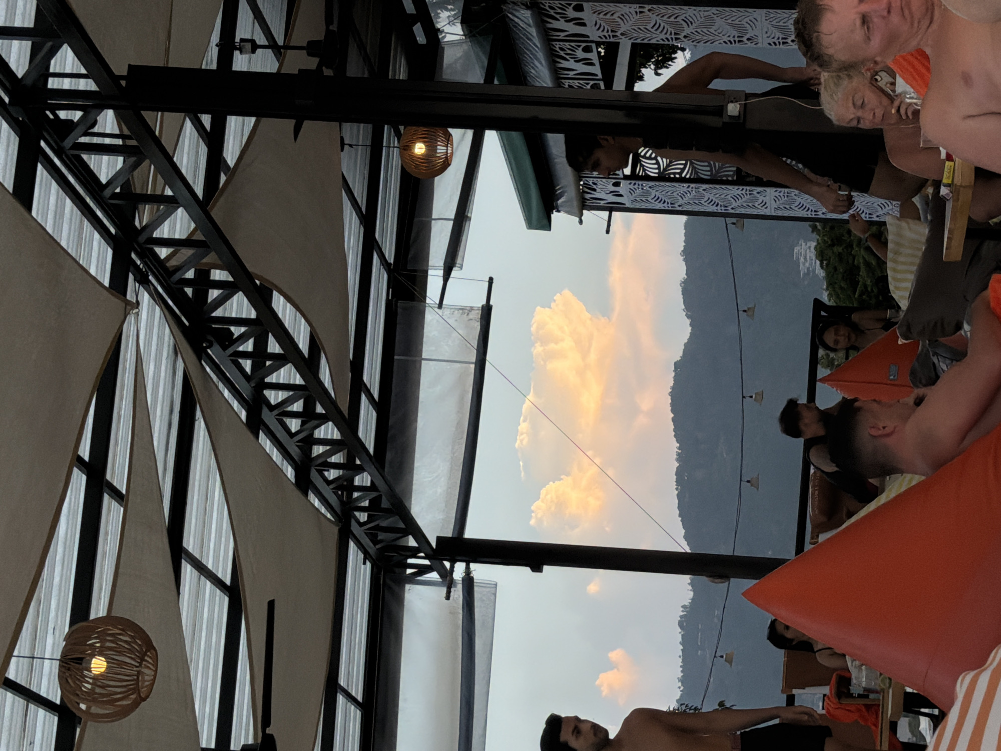 A group of people are gathered under an outdoor canopy at dusk. The canopy is constructed with a metal frame and fabric covers, with some fabric draped and some stretched taut. There are pendant lights hanging from the ceiling. In the foreground, there are large orange beanbag chairs and other seating. In the middle ground, several individuals are visible, some seated and some standing. One man, partially visible on the left, is shirtless. Another man in a black shirt is standing on the right. A woman with blonde hair is speaking on a cell phone, and a man with blonde hair is in the immediate foreground. In the background, there is a mountainous landscape with some buildings visible on the slopes, and a dramatic sky with large, illuminated clouds. The location is likely a resort or restaurant in Ko Samui District, Thailand, given the tropical vegetation and the architectural style.