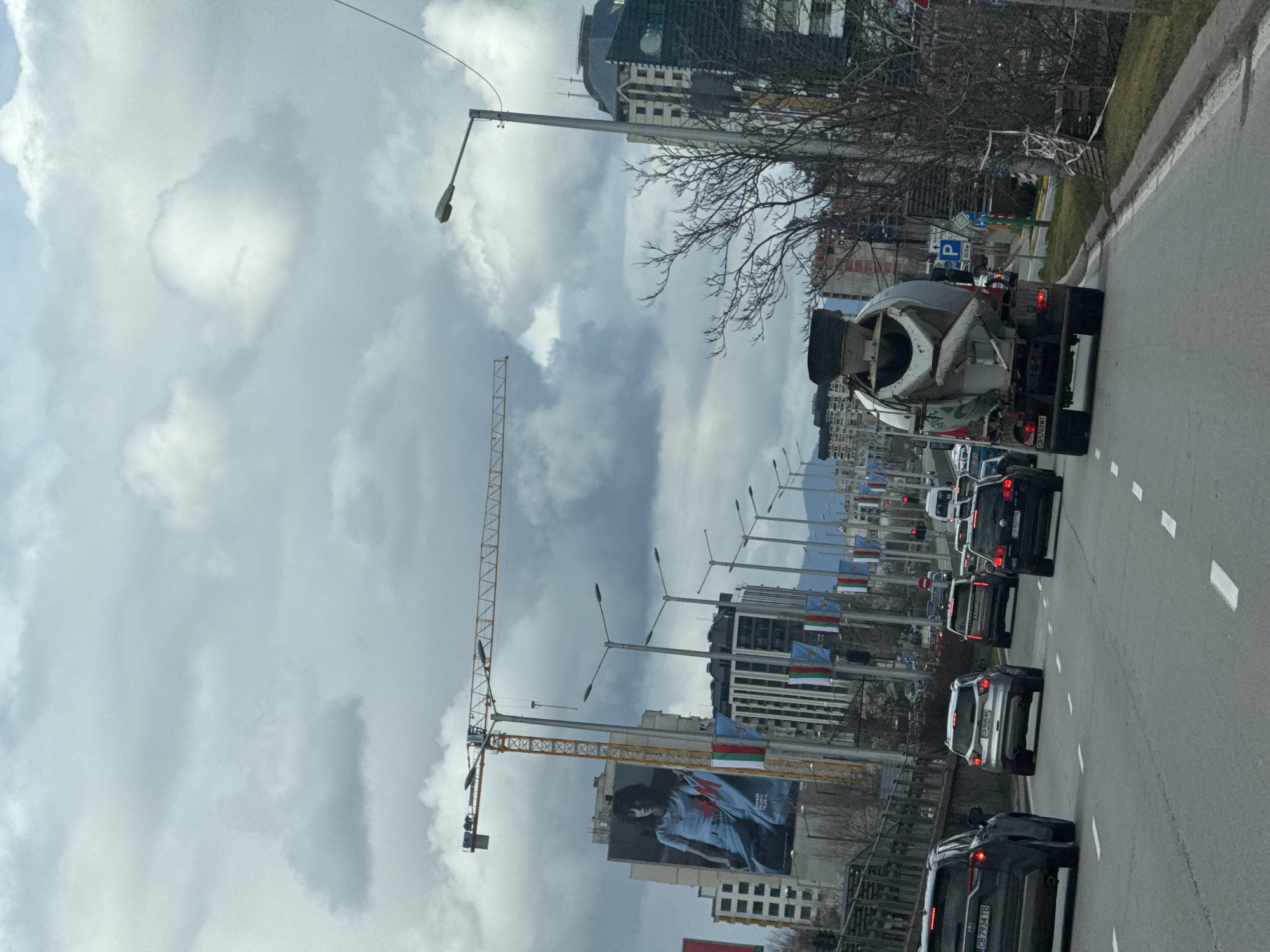 The image depicts a street scene in Sofia, Bulgaria. A line of cars is stopped on the right side of a two-lane road, including a cement truck at the front. Buildings line the left side of the street, with a large billboard visible on one building. A tall crane stands near the buildings. Streetlights are visible along the road. Bulgarian flags are displayed along the street. Mountains are visible in the distance. The sky is overcast with grey clouds. The location appears to be along Tsarigradsko Shose boulevard, facing towards the city center.