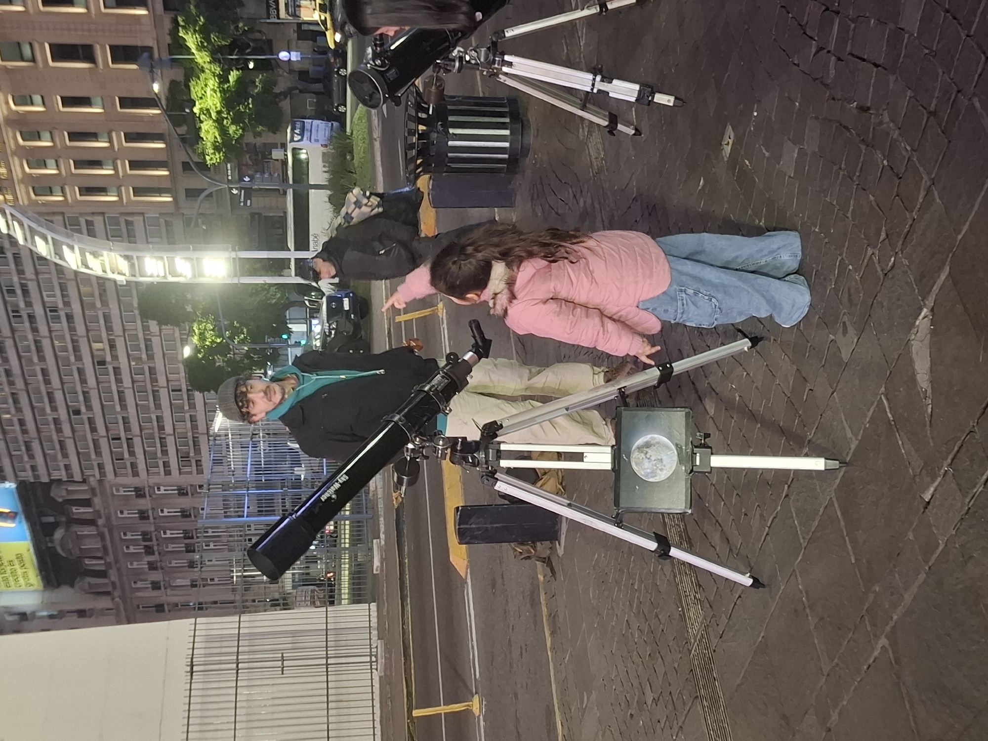 The image depicts a street scene at night in Buenos Aires, Argentina. Two telescopes on tripods are positioned on the paved sidewalk. A man in a black jacket, tan pants, and gray beanie is kneeling and looking at one of the telescopes. Nearby, a young girl in a pink jacket and jeans is kneeling on the sidewalk and pointing, presumably at something in the sky. Another person in a dark jacket is partially visible behind them.
The telescopes are aimed towards the sky. A board with a picture of the moon is attached to one of the tripods. In the background, there are tall buildings with multiple lit windows, indicating an urban environment. Traffic and streetlights are visible on the street.