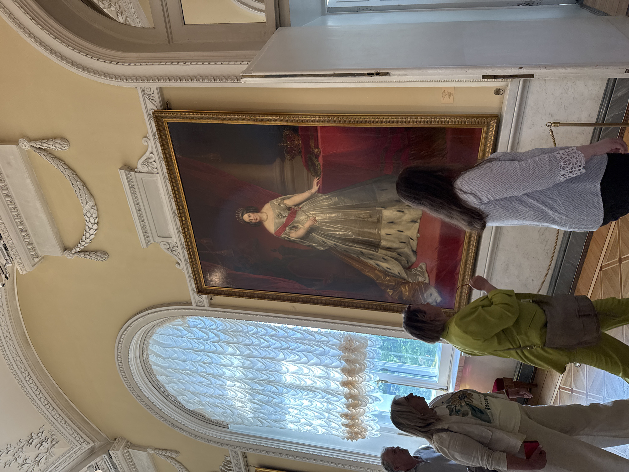 The image depicts a group of people in a museum in Saint Petersburg, Russia, viewing a large portrait. The portrait is of a woman wearing a crown, a red sash, and a formal gown with a fur-trimmed cape, standing next to a table with a crown on it. The room has high ceilings with ornate moldings and a large, arched window with sheer curtains. The walls are painted a pale yellow. The people are casually dressed and appear to be tourists. One woman in a green suit is looking at the painting, while another woman in a cream-colored jacket and white t-shirt is also observing the artwork. A man in a light-colored shirt is partially visible on the left, and another woman in a patterned shirt is in the foreground, facing away from the camera and looking at the portrait. The floor is wooden with an inlaid geometric pattern. A velvet rope barrier is present in front of the painting, separating it from the viewers. The overall impression is a museum visit in an opulent setting.