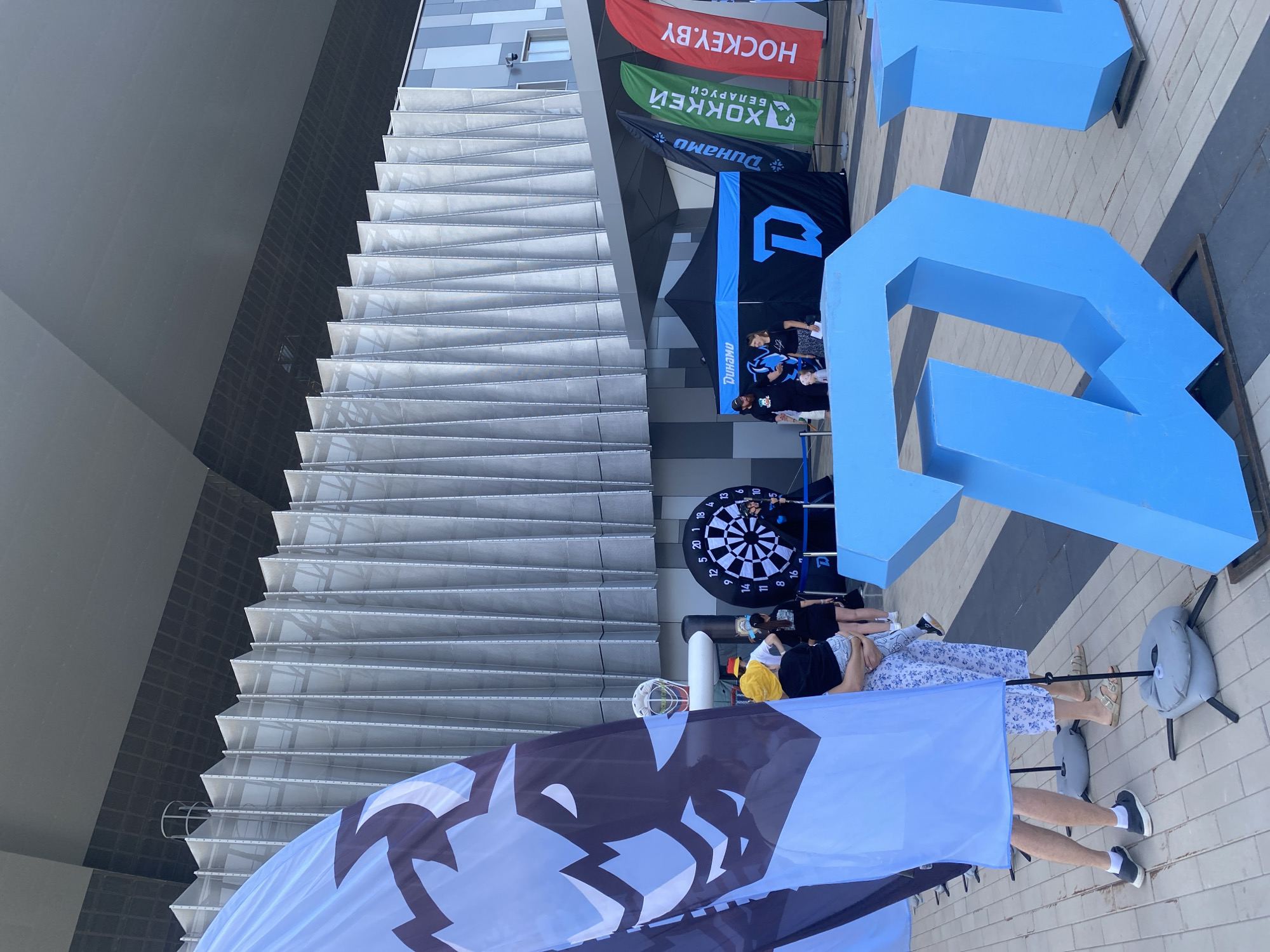 The image depicts an outdoor event area in Minsk, Belarus, in front of a modern building with a distinctive ribbed facade. In the foreground, large, bright blue, three-dimensional letters, seemingly spelling "DW", are prominently displayed on a paved ground surface.
To the left, a large blue flag with a black and white graphic of a stylized wolf's head is unfurled. Behind the flag, several people are gathered. A woman in a patterned dress holds a young child, while another person in a yellow hat stands nearby.
Further back, a black tent with blue lettering and a large blue "D" logo is set up. Two individuals, a man and a woman, stand near the tent, interacting with a child who is wearing a yellow hat. Adjacent to the tent, a large dartboard is set up, and someone is about to throw a dart.
Several vertical flags are displayed to the right of the tent. One flag is green with white Cyrillic lettering that reads "ХОККЕЙ БЕЛАРУСИ" which translates to "HOCKEY BELARUS". Another flag is red with white lettering that reads "HOCKEY.BY". There is also a flag with the "Динамо" logo. The presence of the "HOCKEY.BY" flag and "HOCKEY BELARUS" strongly suggests the event is related to ice hockey and possibly the Belarusian national team or a hockey club. The "Динамо" logo is also often associated with sports teams.
The overall scene suggests a public event or promotional activity taking place outdoors, possibly at a sports venue or a public square in Minsk, given the context provided by the flags. The modern architecture of the building in the background indicates a contemporary urban setting.