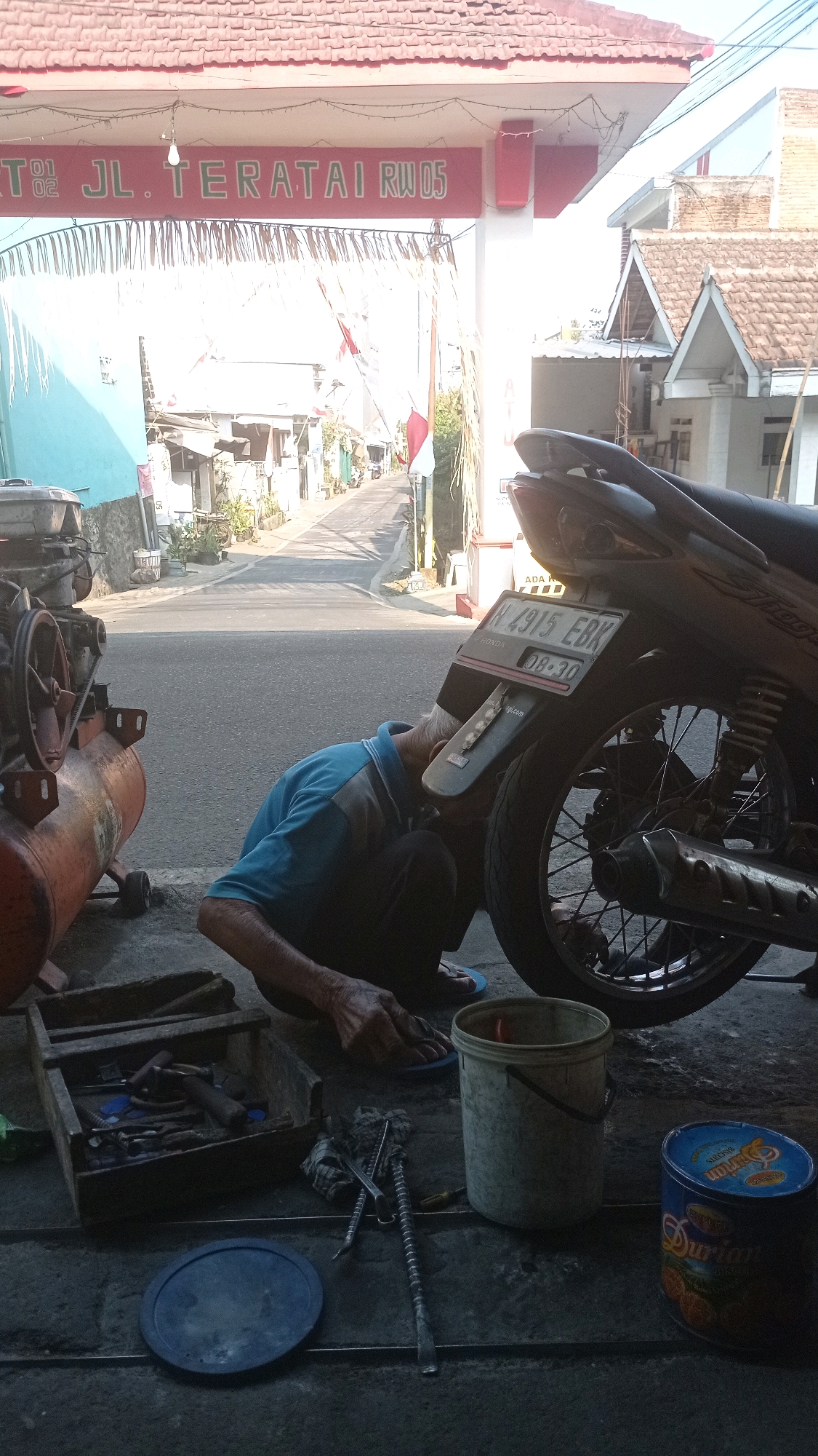 A man is working on a motorcycle at a street-level repair shop in Batu, Indonesia. The shop is under a gate archway that reads "JL. TERATAI RW 05". A blue shirted man is kneeling, bent over the rear wheel of a motorcycle, appearing to be making repairs. Tools are scattered on the ground around him, including a wooden toolbox filled with wrenches and other implements, a blue lid, and a metal bucket. An orange air compressor is also visible to the left of the scene. The street behind the man is paved and lined with houses and small shops, and Indonesian flags are visible, suggesting it may be a patriotic holiday. The lighting suggests it is daytime, possibly late afternoon, as shadows are long.