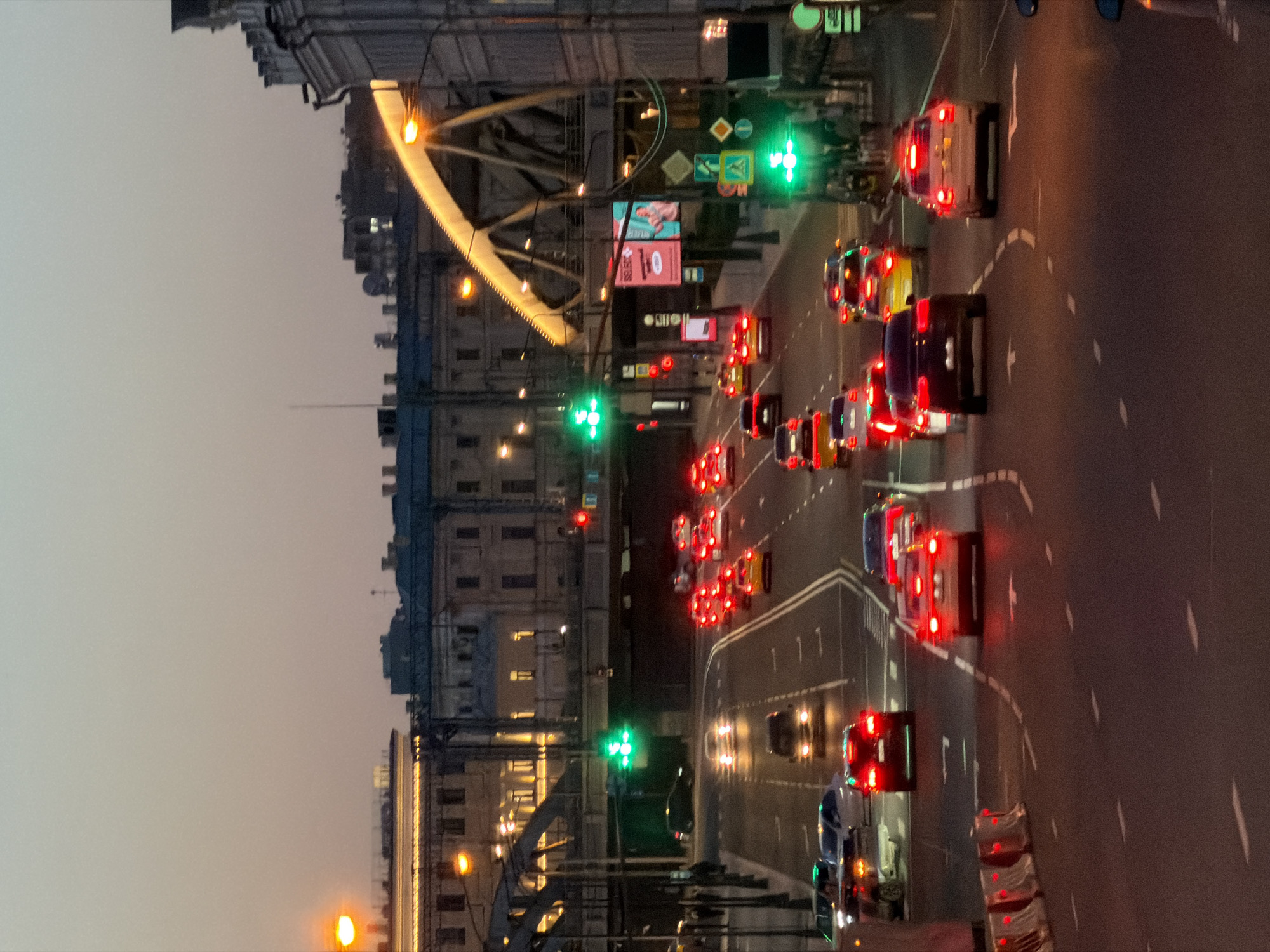 The image depicts a traffic scene on a multi-lane road in Moscow, Russia, at dusk. The road is paved and marked with white dashed lines. Numerous cars, many with their brake lights illuminated, are in motion or stopped at traffic signals. Several vehicles appear to be taxis, identified by their yellow color. Traffic lights, showing green, are visible at intersections. Overhead, a bridge structure with illuminated arches spans across the road, and adjacent buildings with illuminated windows are visible. Streetlights and building lights cast a warm glow on the scene. The sky is a pale, dusky hue, indicating either dawn or twilight. The overall atmosphere is that of a busy urban environment transitioning from day to night.