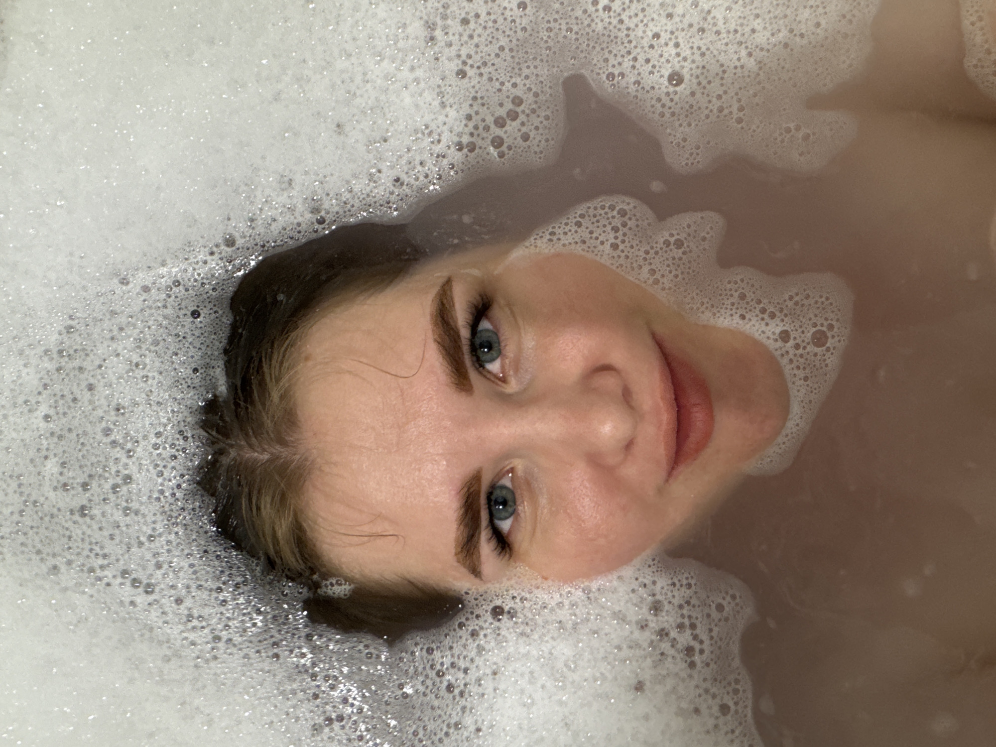 The image displays a close-up, high-angle view of a young woman's face as she lies submerged in a bathtub filled with foamy water. Her head is partially underwater, with her wet, light brown hair blending into the surrounding bubbles. She gazes directly upwards at the camera with a gentle, relaxed expression and a subtle smile. Her prominent blue-grey eyes are wide open, framed by distinct eyelashes and well-groomed eyebrows.
The bathwater is largely covered by a thick layer of bright white foam, which dominates much of the frame, especially the upper and left sections. Beneath the bubbles, the visible bathwater has a muted, reddish-brown or pinkish-beige hue. The contrast between the bright foam, the water, and her fair skin creates visual depth. The scene suggests an indoor setting, likely a bathroom, contributing to a sense of privacy and relaxation. There is no visible text in the image. The location context provided is Ферма, Russia.