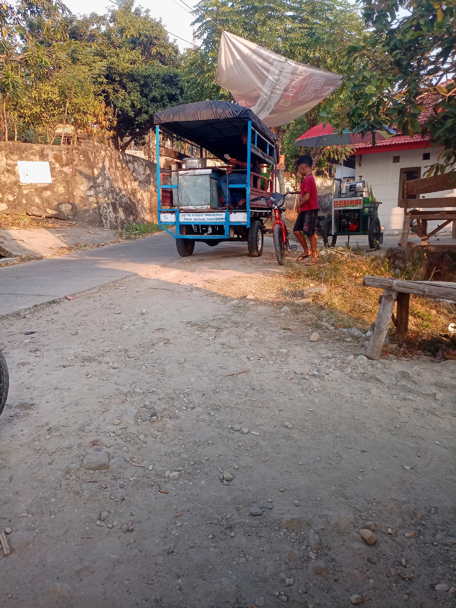A dirt road is bordered by a stone wall and vegetation on the left, and a building and a wooden bench on the right. A three-wheeled vehicle, likely a modified motorcycle with a cargo area covered by a tarp, is parked on the road. Inside the cargo area, there appear to be coolers or storage containers. A young boy in a red shirt and dark shorts stands near the vehicle. To the right of the vehicle, a sugarcane juicing machine is visible. The scene is illuminated by natural light, suggesting it is daytime. The presence of the sugarcane juicer and the overall setting suggests this may be a street vendor in a rural or semi-urban area of Manggar, Indonesia. The writing on the vehicle and sugarcane machine is in Indonesian. The text on the vehicle reads "KU TAK BERHENTI MENCINTAINYA, HANYA BERHENTI MENUNJUKKANNYA" which translates to "I won't stop loving it, I'll only stop showing it." The sugarcane machine has the text "ESTEBU HIJAU" which means "Green Sugarcane Juice."