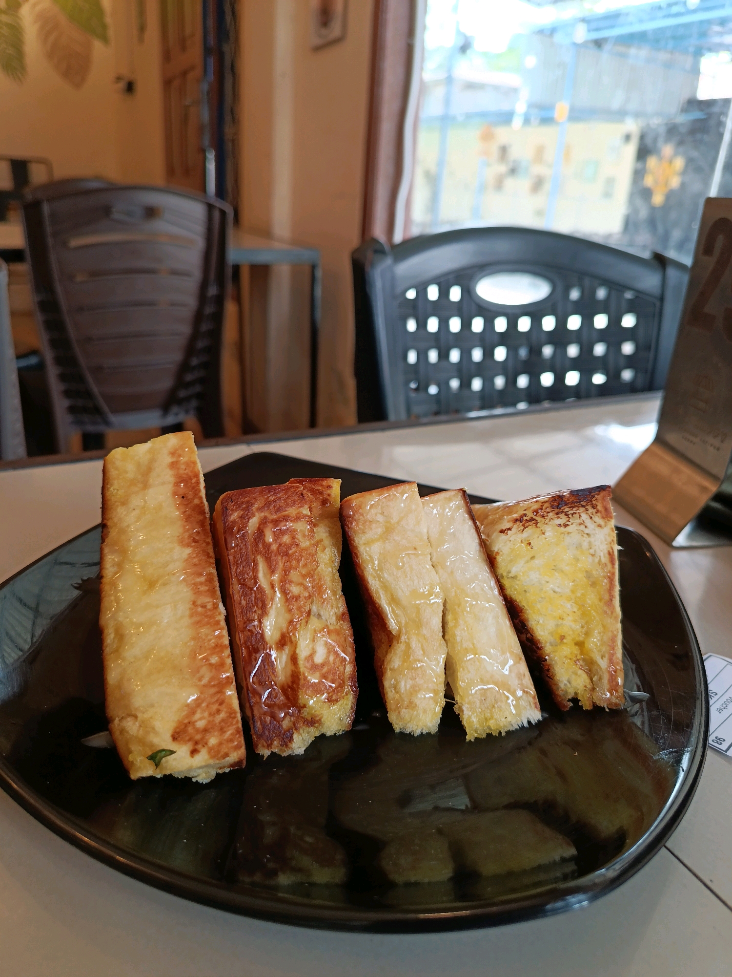 Four pieces of toast are arranged on a black plate. The toast pieces are rectangular and appear to be sweet or savory, with a glossy coating. The toast on the far left has a light brown, slightly crispy exterior. The second piece from the left is more uniformly golden brown. The third and fourth pieces from the left are lighter in color, with a more porous texture. The toast appears to be drizzled with a light-colored syrup or sauce. In the background, the image shows chairs and a window, suggesting an indoor seating area, possibly a cafe or restaurant. The lighting is natural and comes from the window, creating some reflections on the plate.