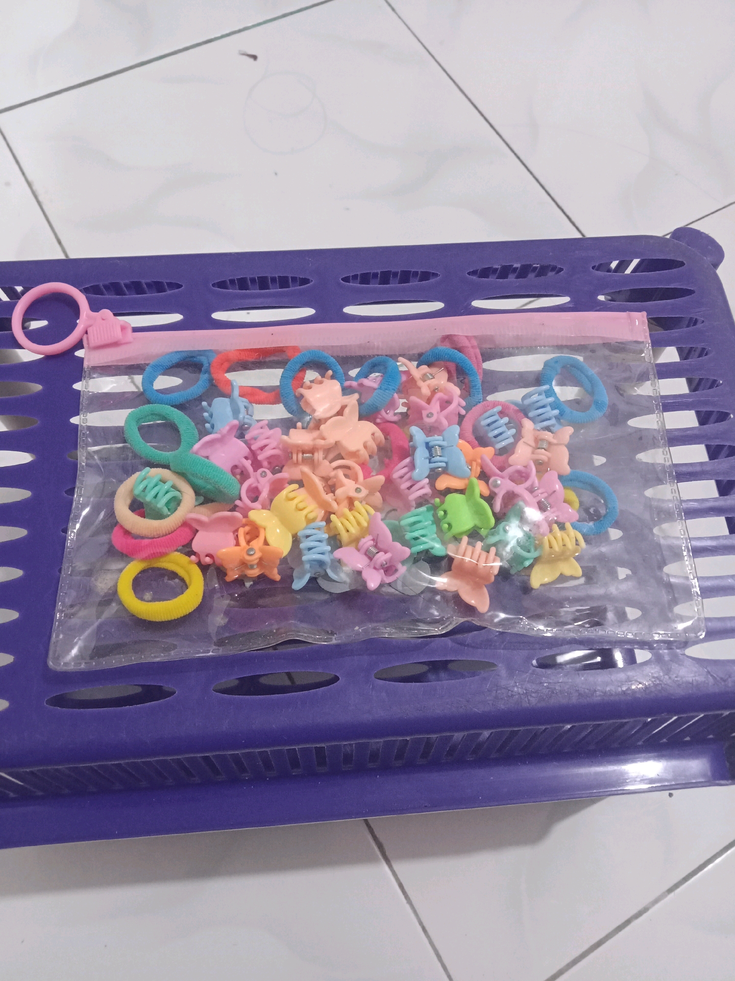 A transparent zippered pouch filled with colorful hair accessories, including small claw clips and elastic hair ties, rests on a purple plastic grate-like surface. The pouch has a pink zipper and a pink plastic ring pull. The hair accessories are in shades of pink, blue, yellow, orange, and green. The purple grate is placed on a white tiled floor with faint lines. The overall scene suggests a child's personal belongings or a storage solution for small items.