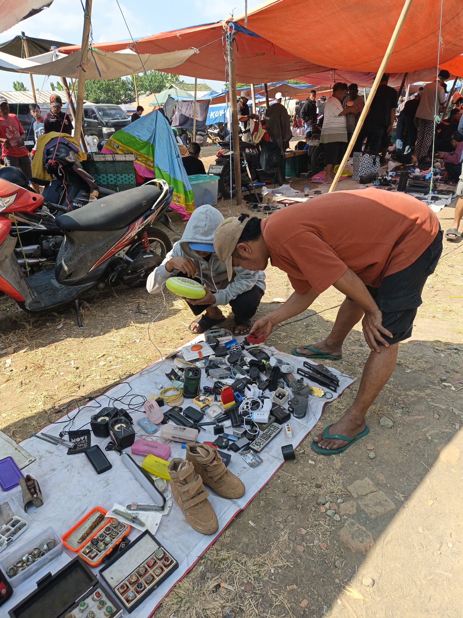 In Jatinom, Indonesia, an outdoor market scene unfolds under an orange tarpaulin canopy supported by wooden poles. In the foreground, two individuals are examining assorted electronic items and accessories laid out on a white sheet. One person, wearing a gray hoodie and a baseball cap, holds a yellow device. The other, in an orange t-shirt and black shorts, points at items on the sheet. A red and black motorcycle is parked behind them. The ground is a mix of dirt and dry grass. In the background, other market stalls and people are visible, along with vehicles. The scene appears to be a bustling marketplace with various goods for sale.