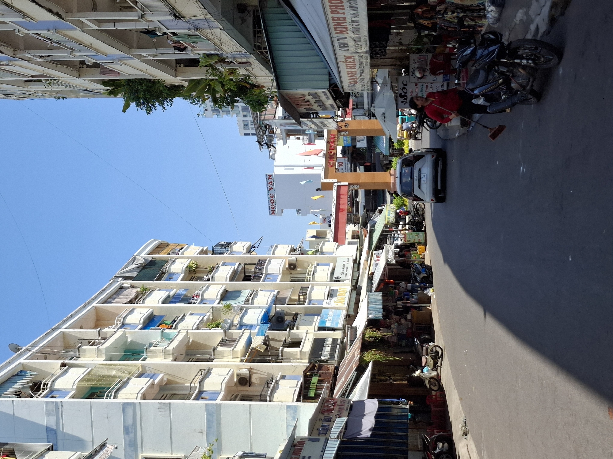 The image shows a street scene in Nha Trang Ward, Vietnam, characterized by a mix of residential buildings and market stalls. On the left, a tall, light-colored apartment building with multiple balconies, each adorned with various shades of blue and yellow awnings, dominates the view. The building's facade features numerous windows and air conditioning units. Below the apartment building, a row of market stalls with colorful awnings lines the street.
On the right side of the street, another apartment building, also light-colored, is visible, with balconies and plants. In front of this building, more market stalls are set up, displaying goods and clothing.
The street itself is paved and appears to be a busy thoroughfare. Several motorbikes are parked along the sides and in the street. A white car is seen driving away from the viewer, and a woman in a red shirt and black pants is sweeping the street with a broom near a parked motorbike. In the background, a large archway with signs in Vietnamese is visible, suggesting this might be an entrance to a market or a specific area. The sky is clear and blue, indicating a sunny day. The overall atmosphere is that of a vibrant urban environment with commercial activity.
