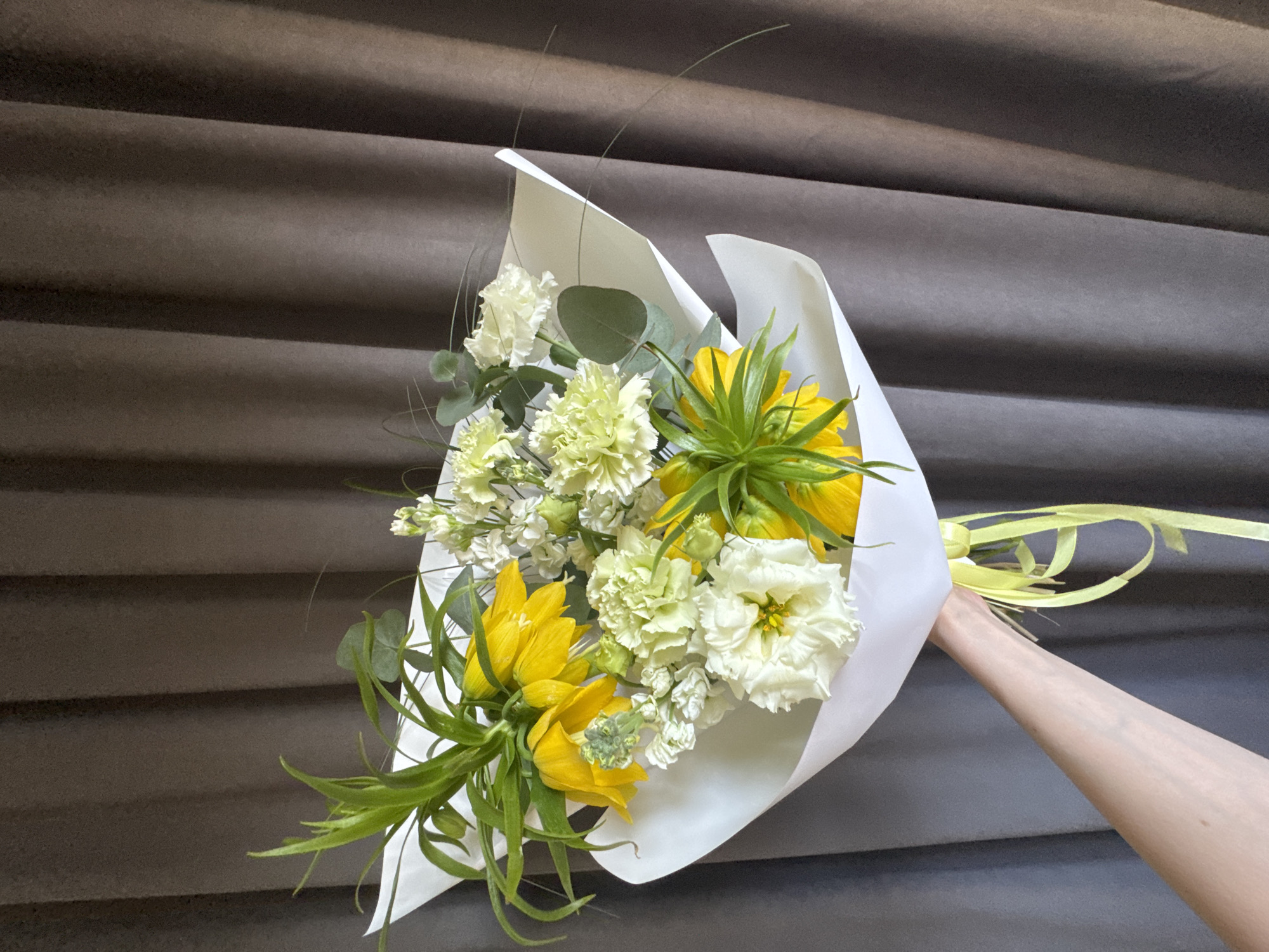 A bouquet of flowers is being held up against a brown, textured background, which is potentially a wall. The bouquet is wrapped in white paper and tied with a yellow ribbon. It contains a mix of flowers, including yellow Fritillaria imperialis, white and green carnations, white lisianthus and white stocks. A hand is holding the bouquet up. There is no indication of this image being in Izhevsk, Russia.